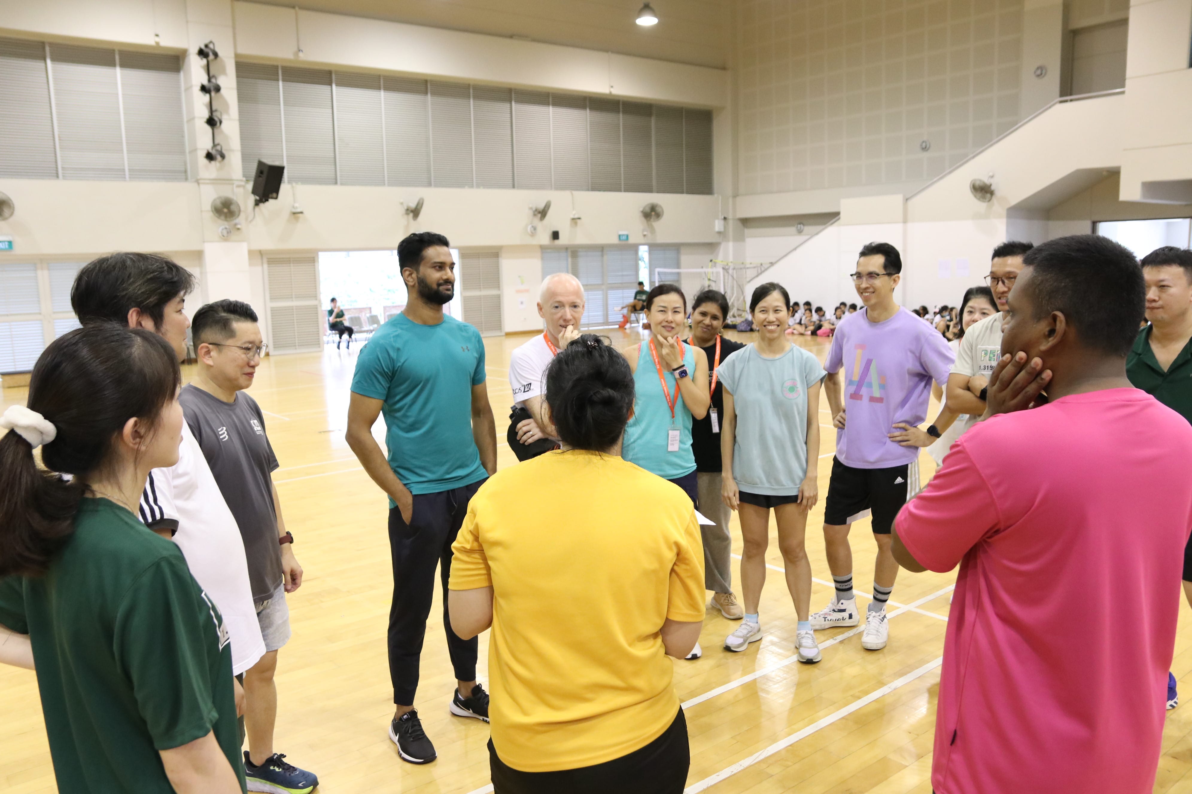 Group of adults in a gym stand in a circle, listening to a person in a yellow shirt facing away.
