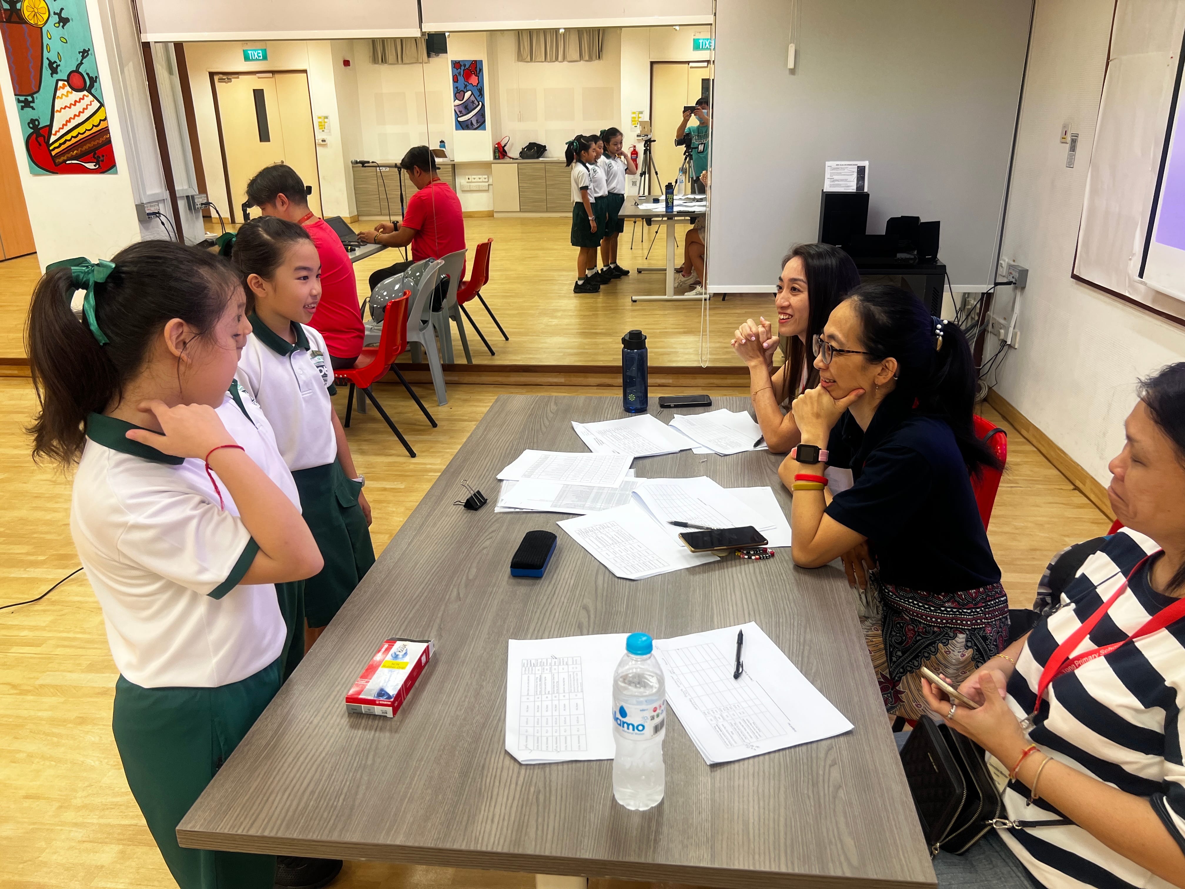 Two schoolgirls in uniforms talk to two women at a table with papers and phones in a hall.
