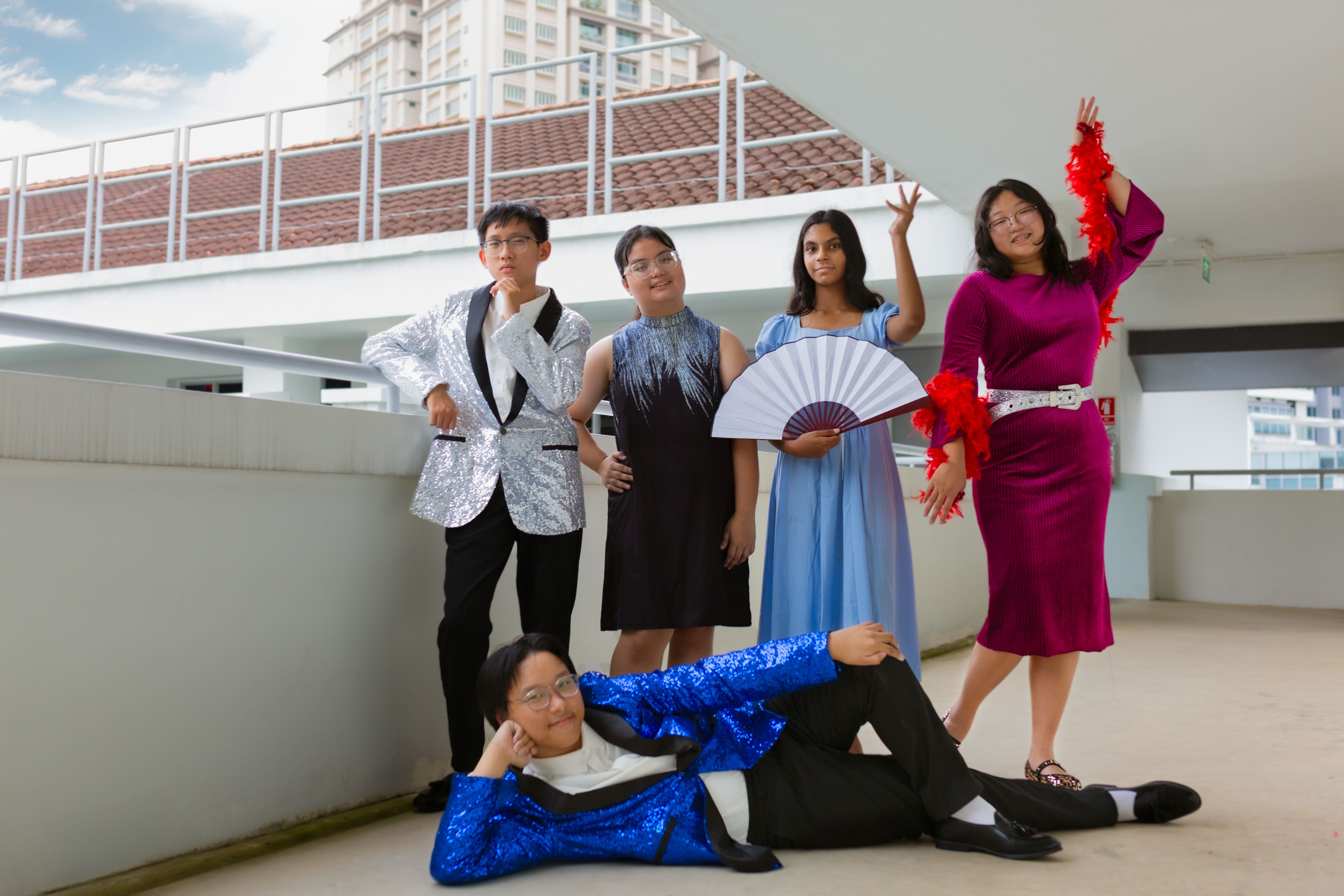 Five teens in glamorous outfits pose on a rooftop, one lying down in a blue sequin jacket, others standing in formal wear.