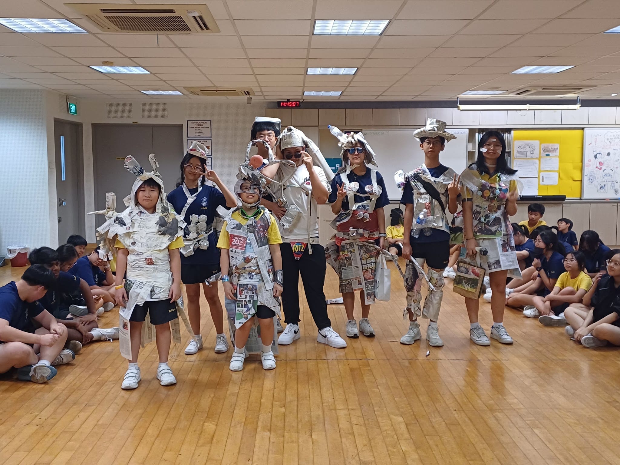 Several children wearing creative costumes made from newspaper and recycled materials stand in a school hall.