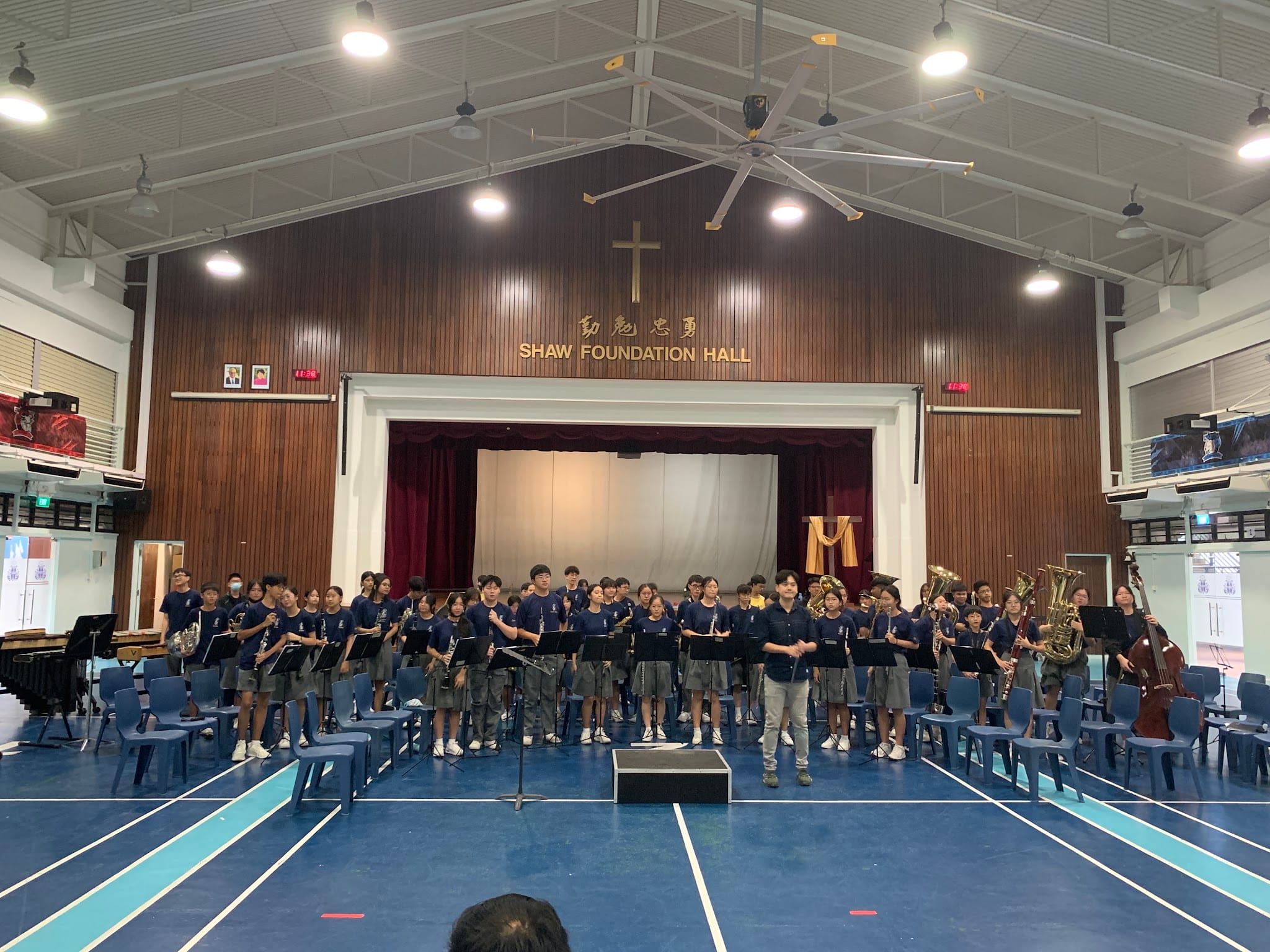 Student band in navy uniforms standing with instruments in Shaw Foundation Hall, led by a conductor on a podium.