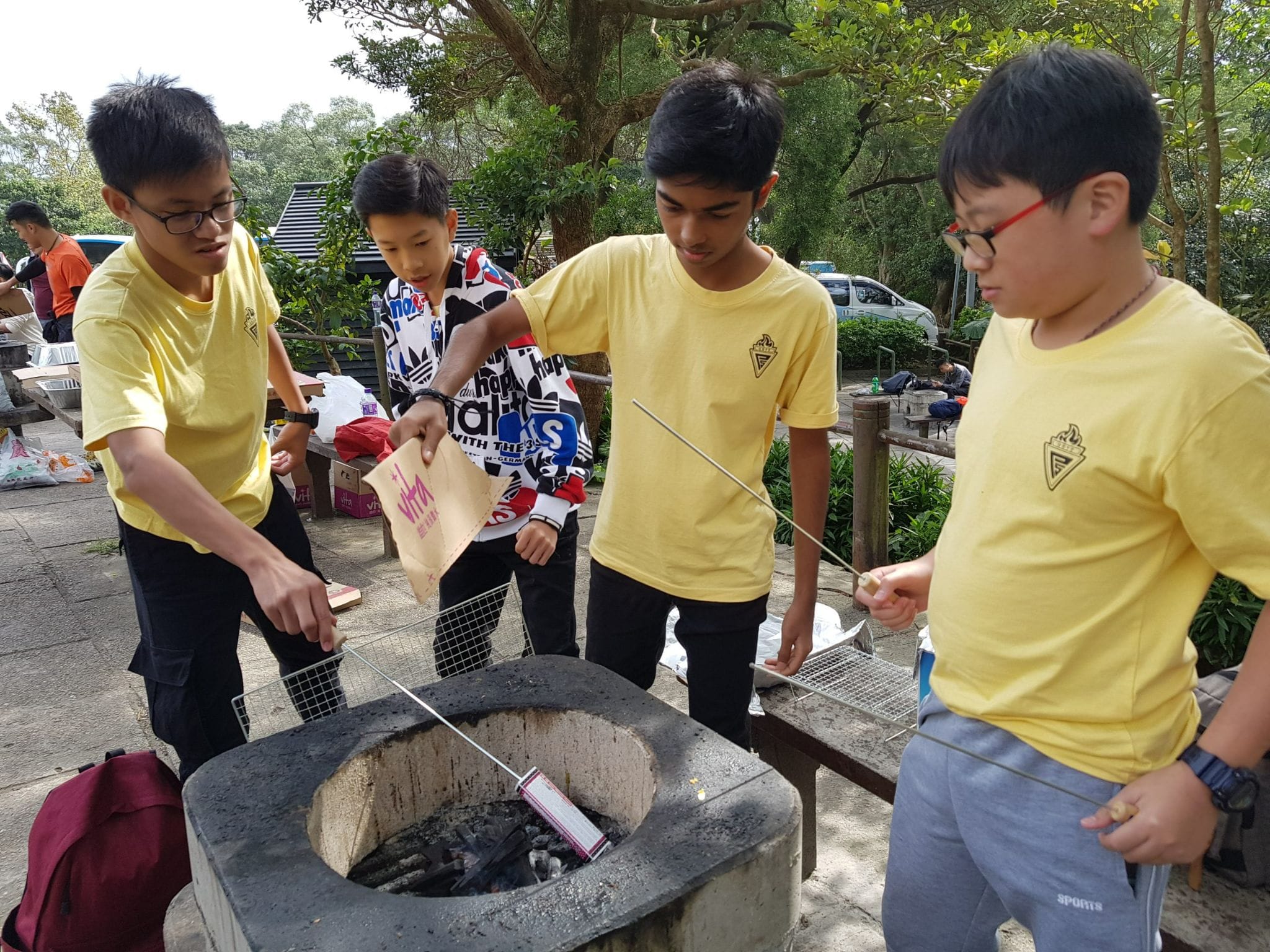 Four boys gathered around an outdoor stone barbecue pit, holding metal skewers and a grill basket over charcoal.