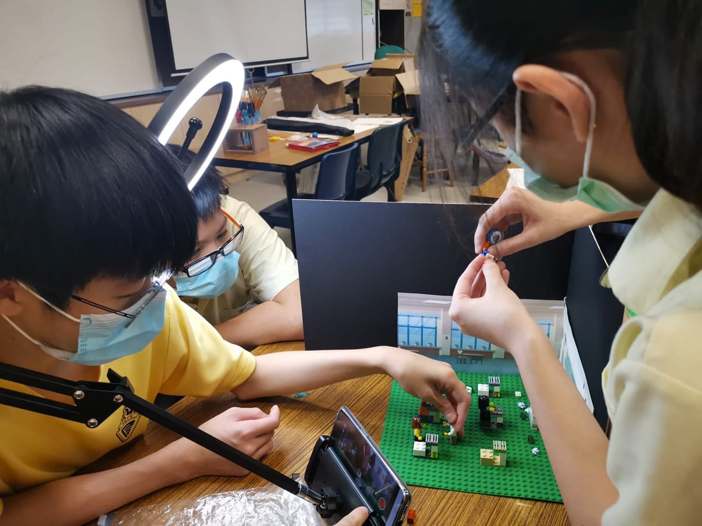 Two masked children build a Lego scene on a green baseplate while recording it with a phone and ring light.