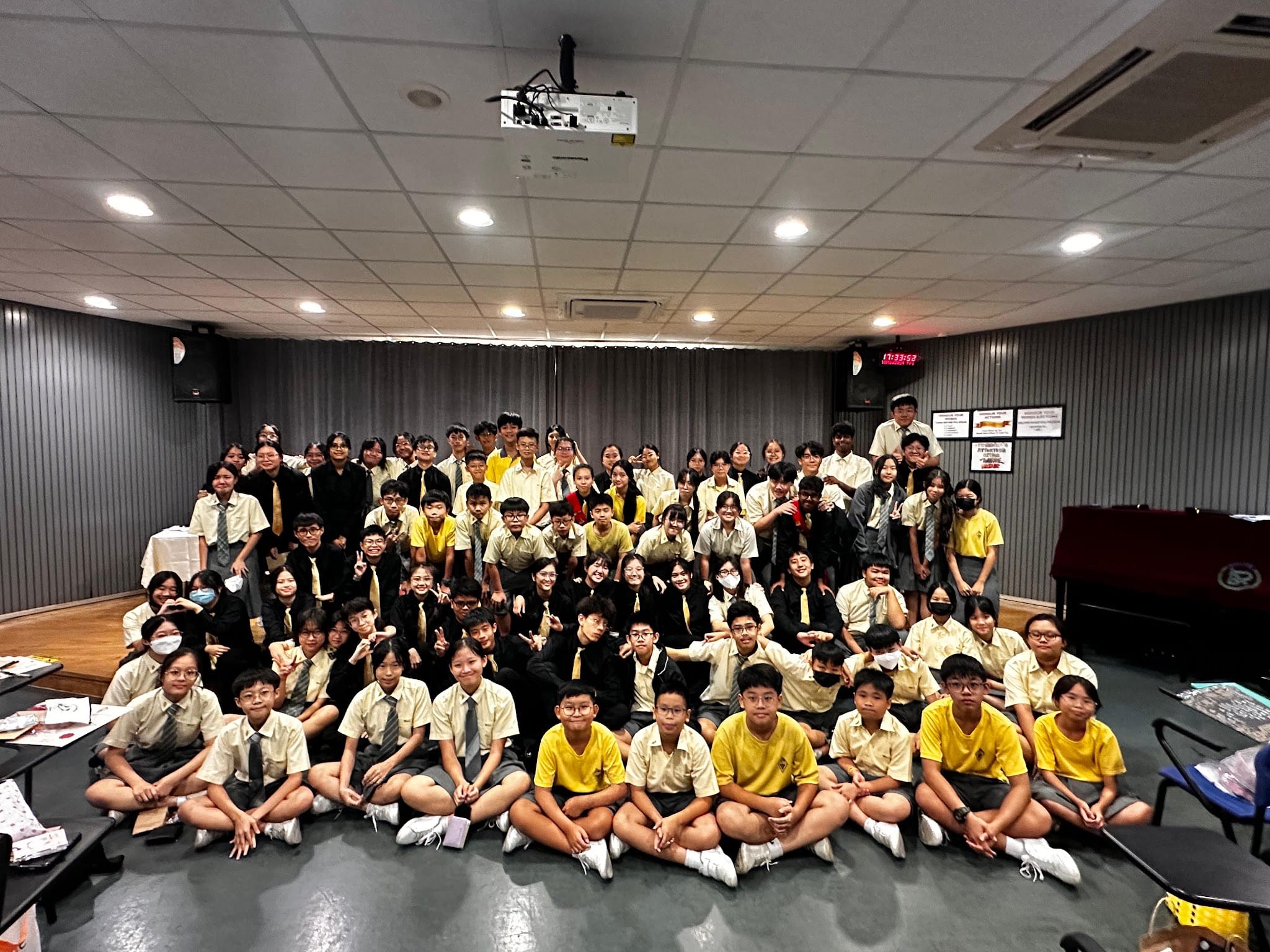 Large group of school students in yellow and black uniforms posing together in an indoor hall for a group photo.