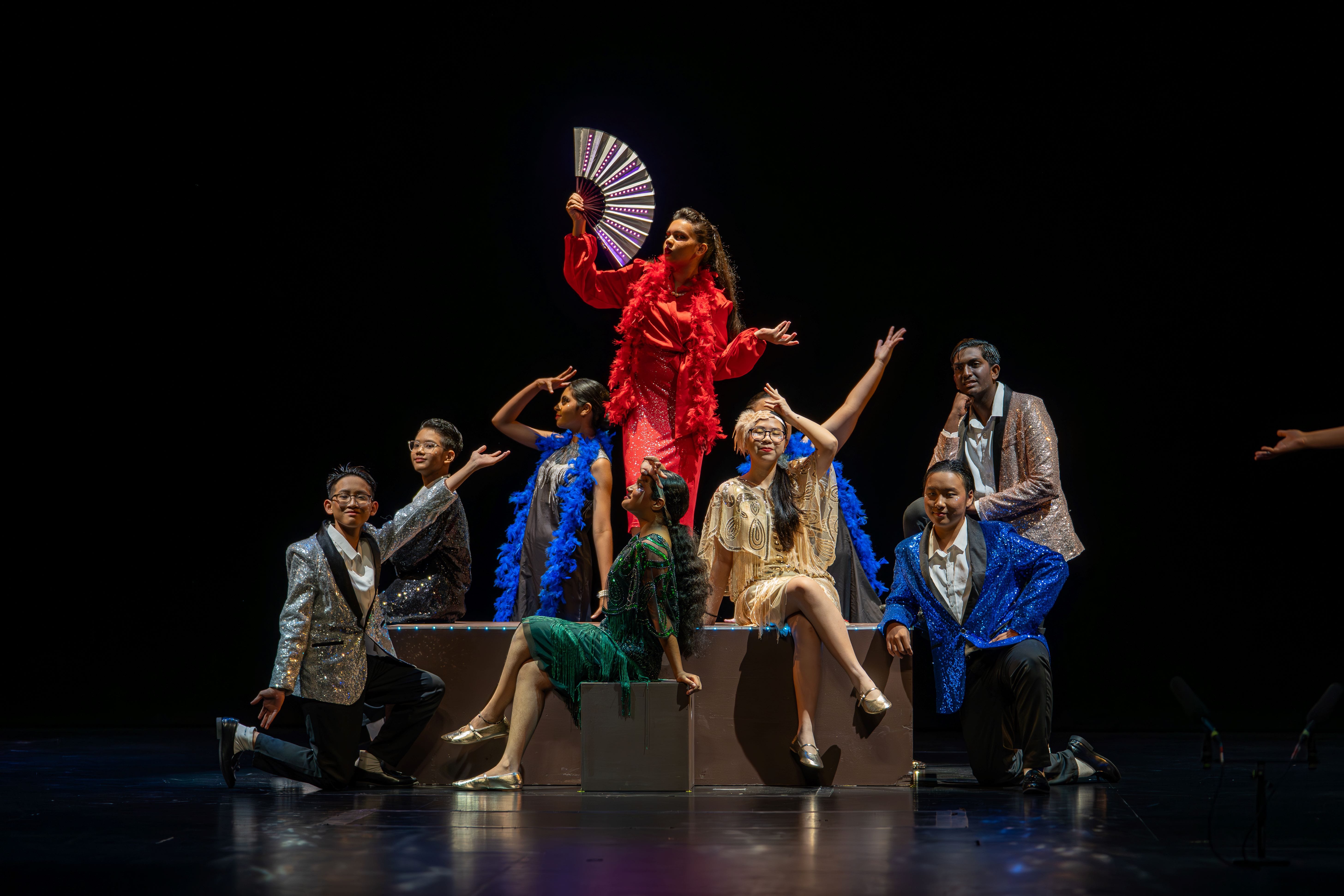 A theatrical cast in sequined and feathered costumes poses on a dark stage, led by a performer in red holding a fan.