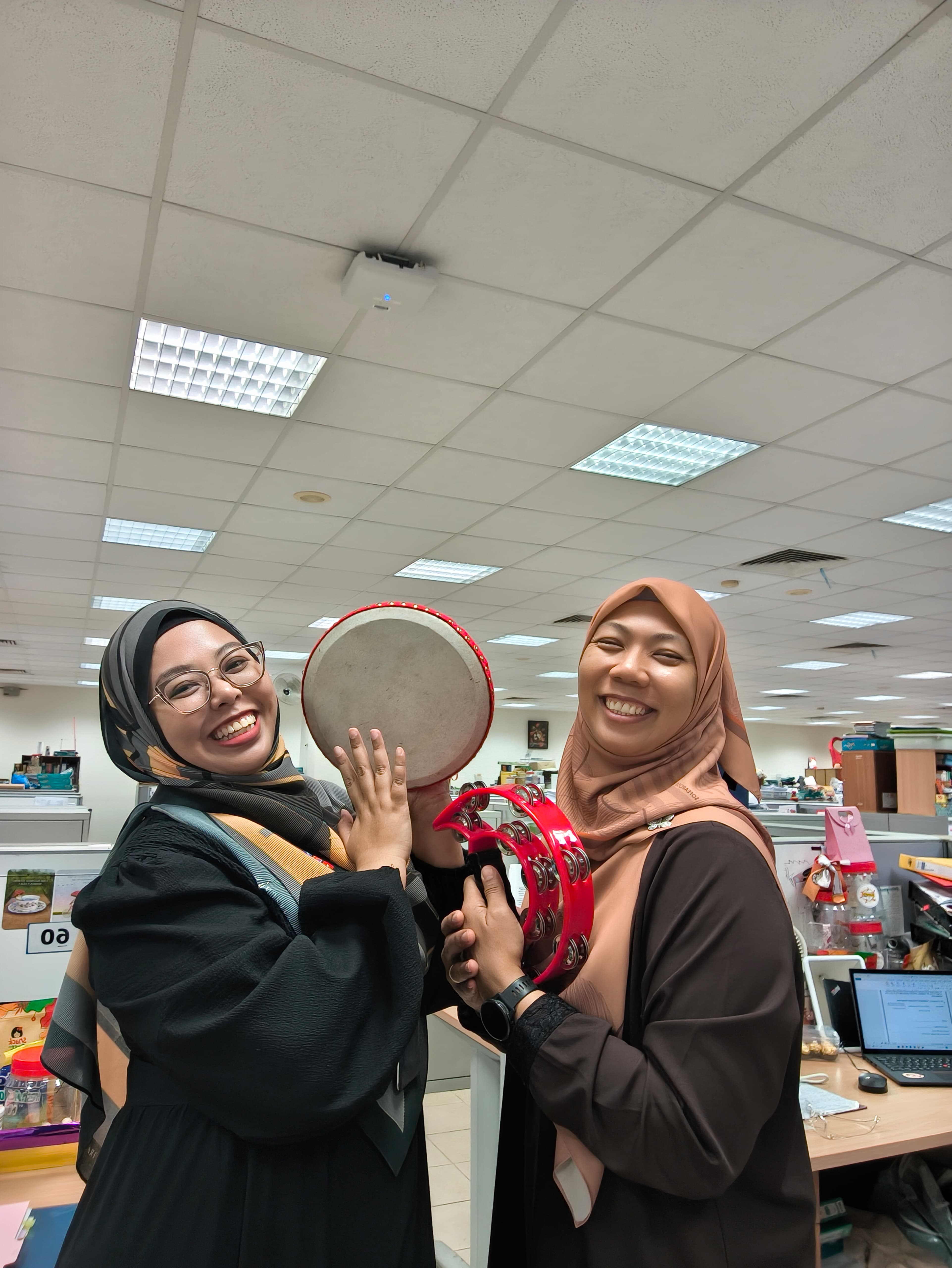 Two smiling women wearing hijabs hold a red tambourine and small drum in a bright office setting.