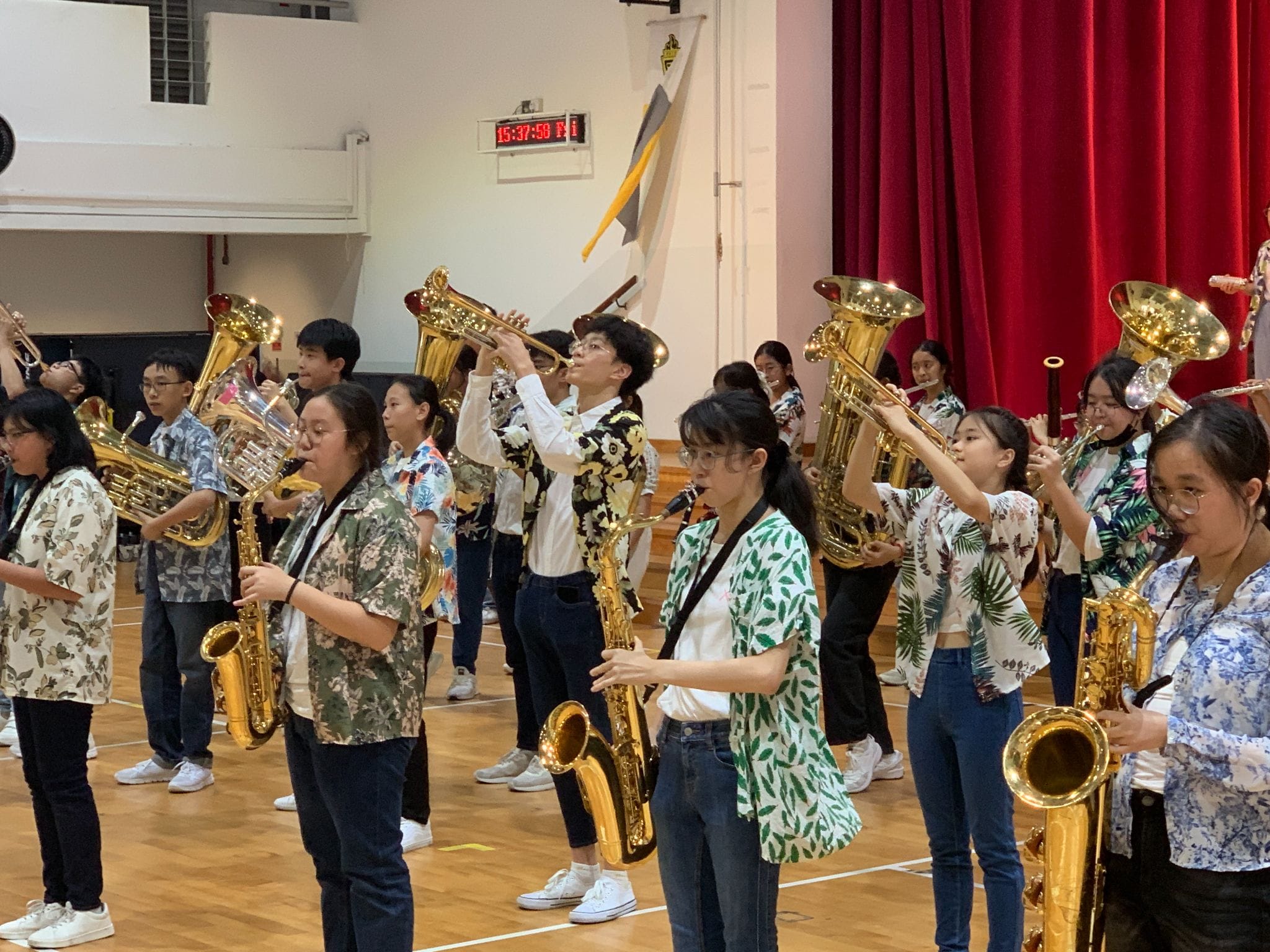 Young students in floral shirts play brass and woodwind instruments in a school hall with a red curtain.