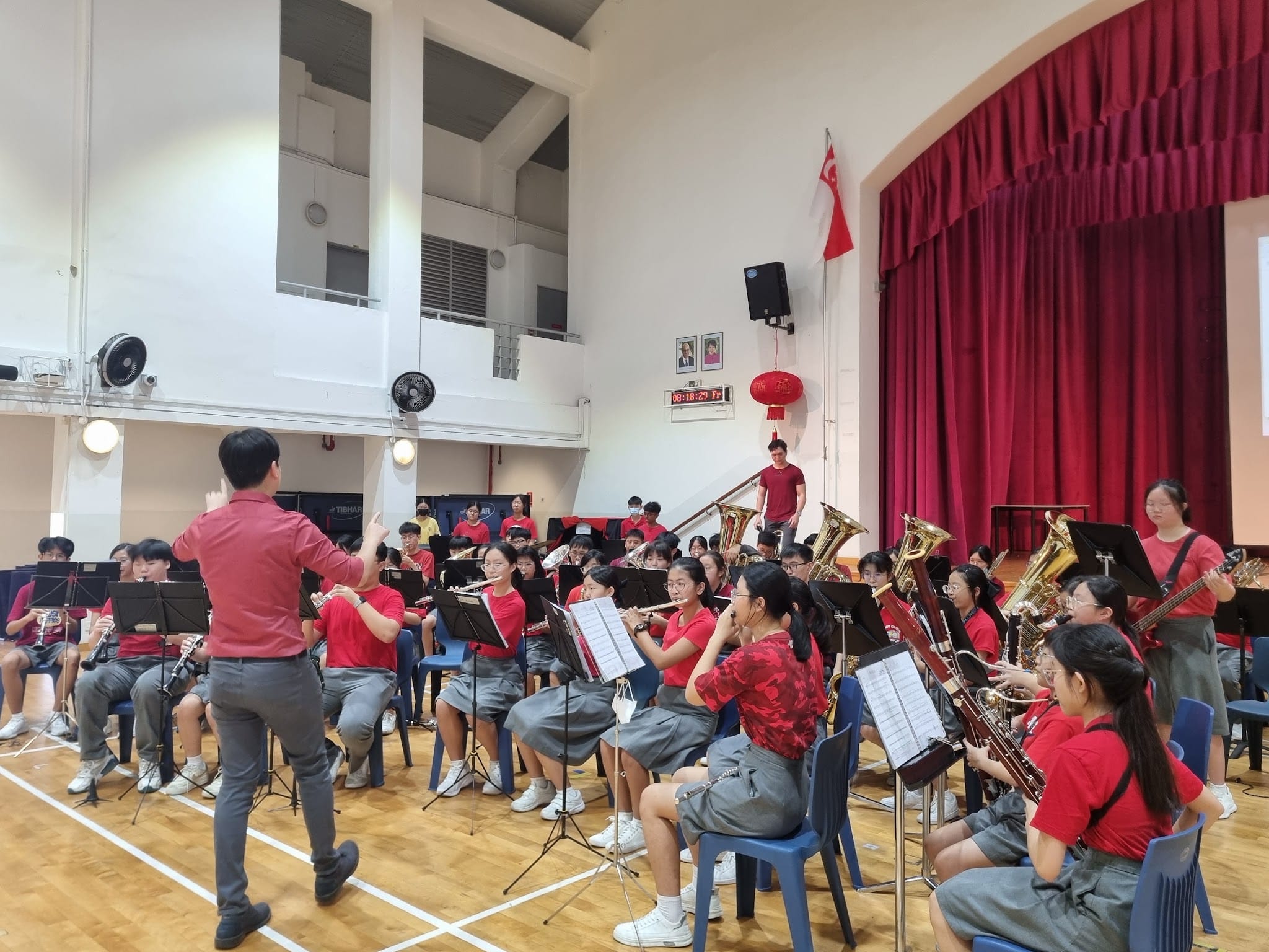 A conductor in red leads a large student band in red shirts playing brass and wind instruments in a school hall.