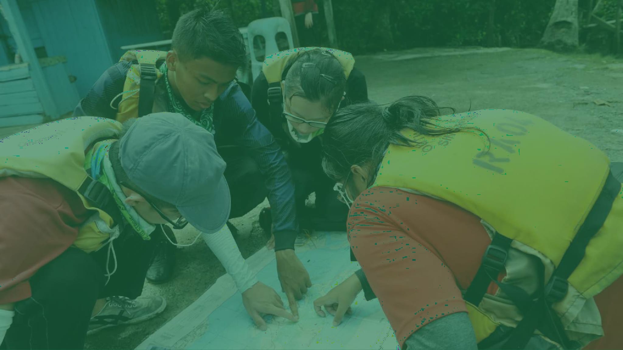 Four people in yellow vests and backpacks lean over a large map on the ground, pointing at it outdoors.