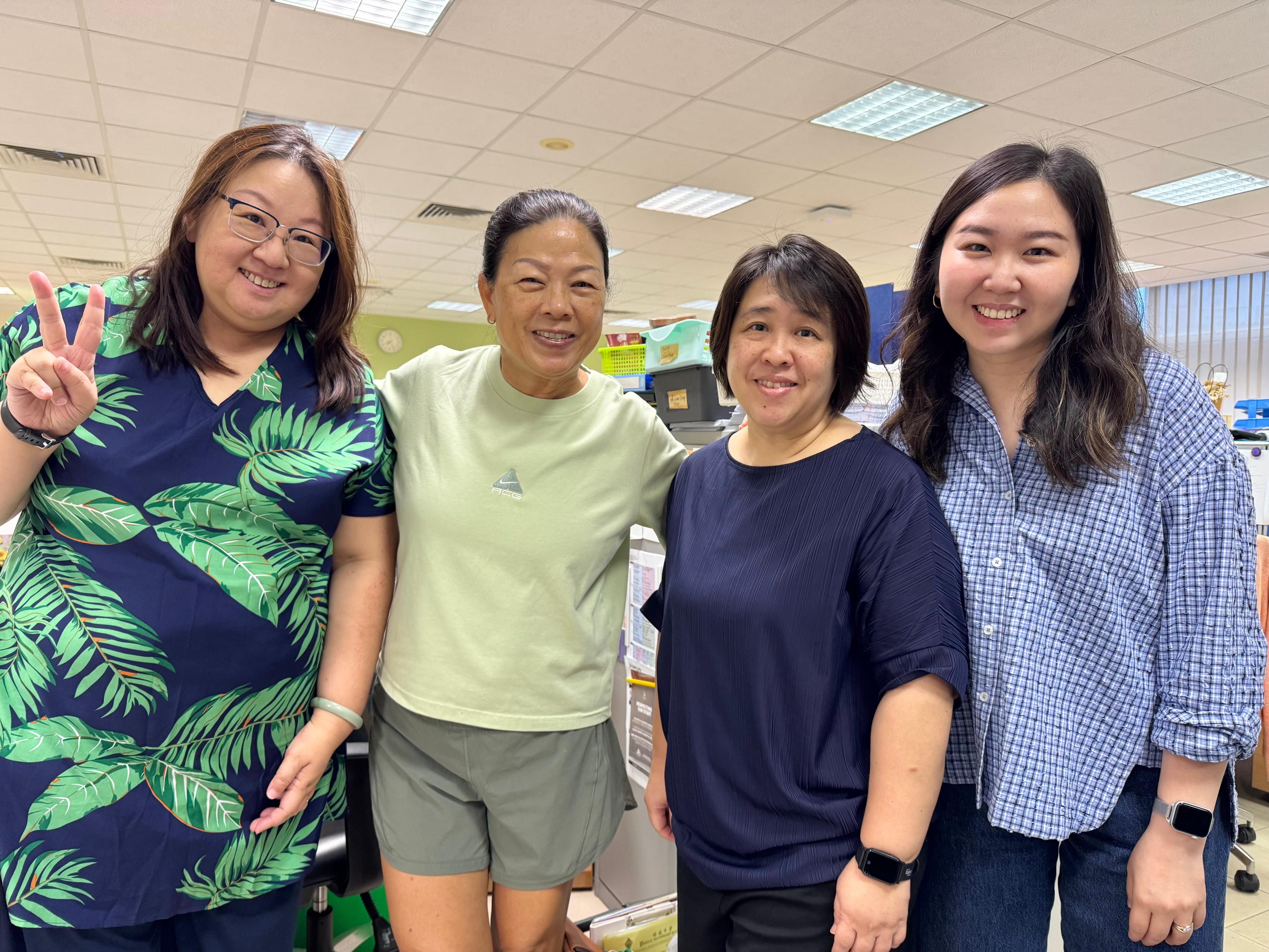 Four smiling women stand together in an office, one making a peace sign, wearing casual colorful tops.