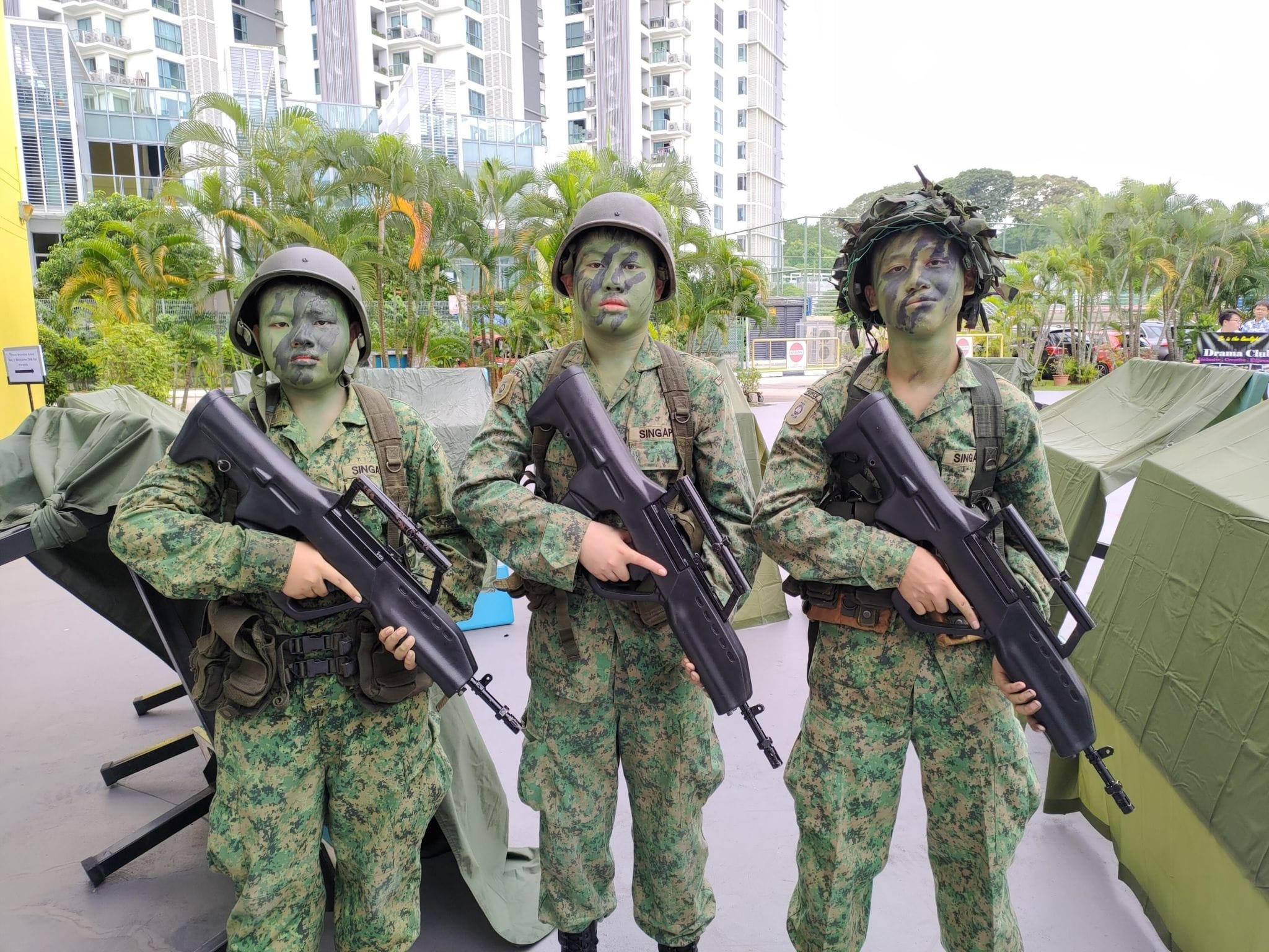 Three soldiers in camouflage uniforms with face paint and helmets holding rifles outdoors near tall buildings.
