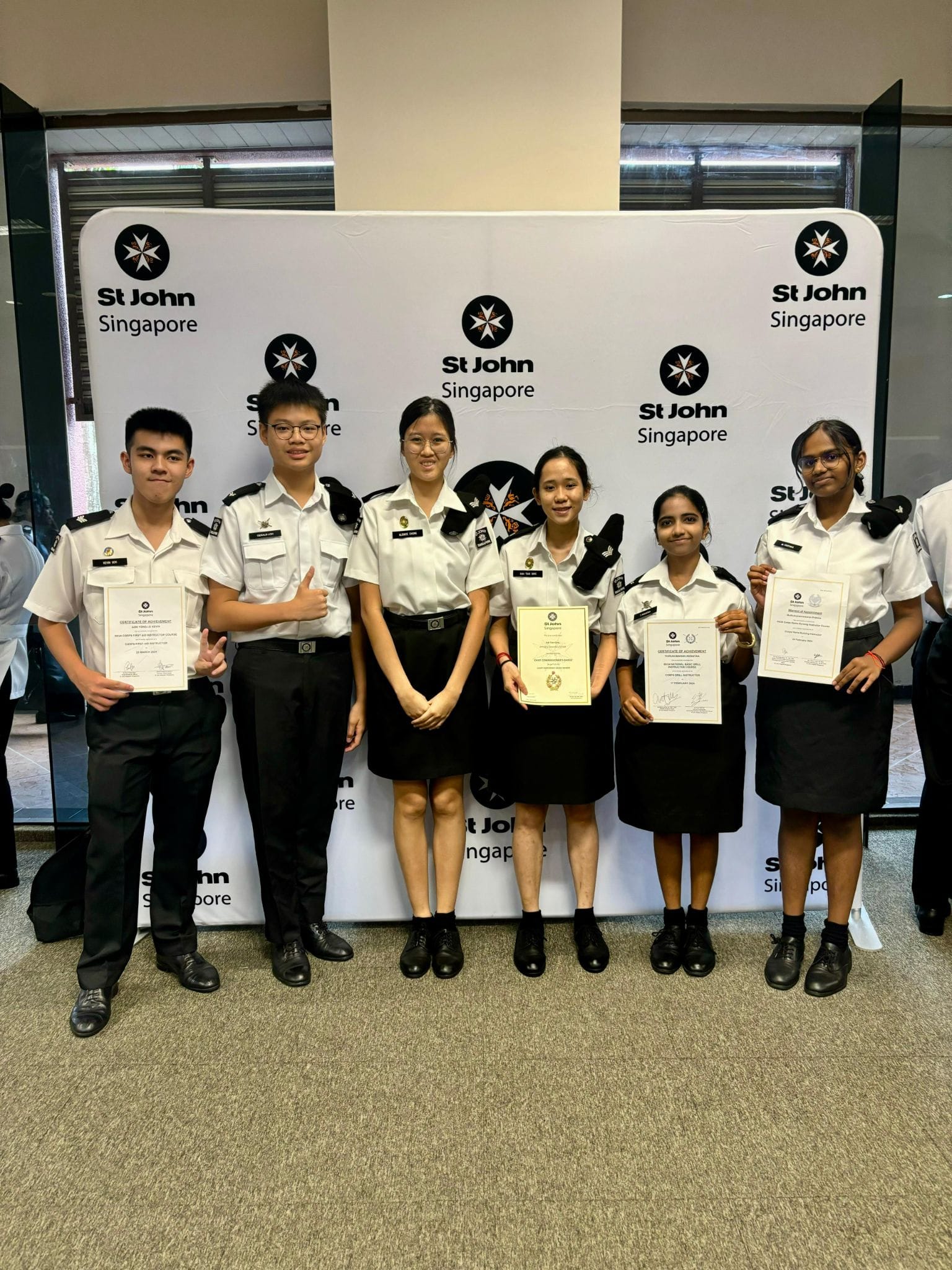 Six young people in St John Singapore uniforms pose with certificates in front of a St John Singapore backdrop.