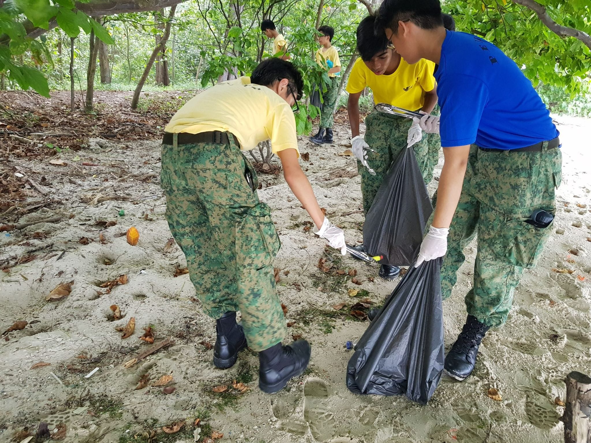 Youth in camouflage pants and gloves collecting litter into a black garbage bag on a sandy beach area.