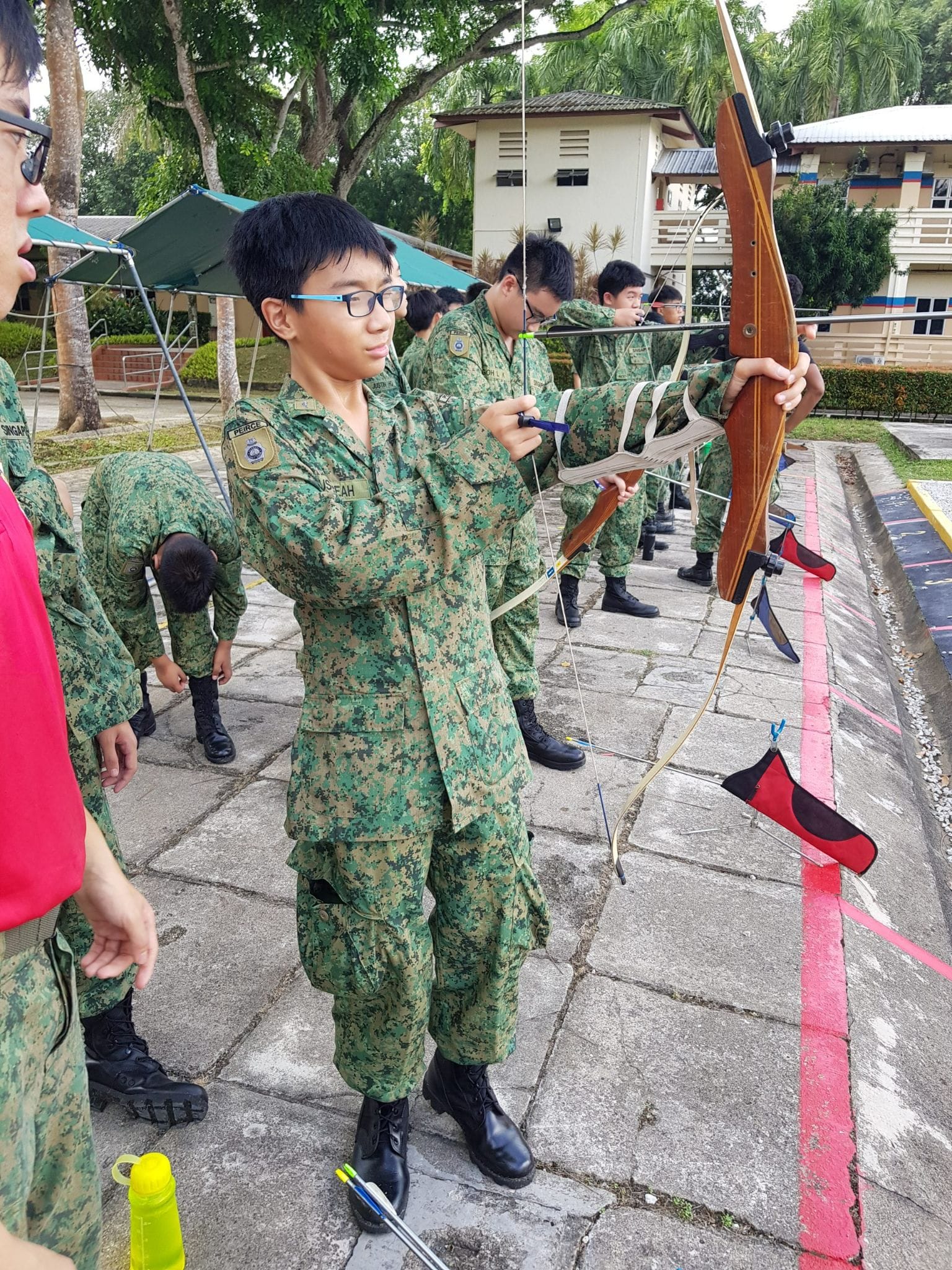Youth in camouflage uniforms practicing archery outdoors, with one boy in the foreground drawing a wooden bow.