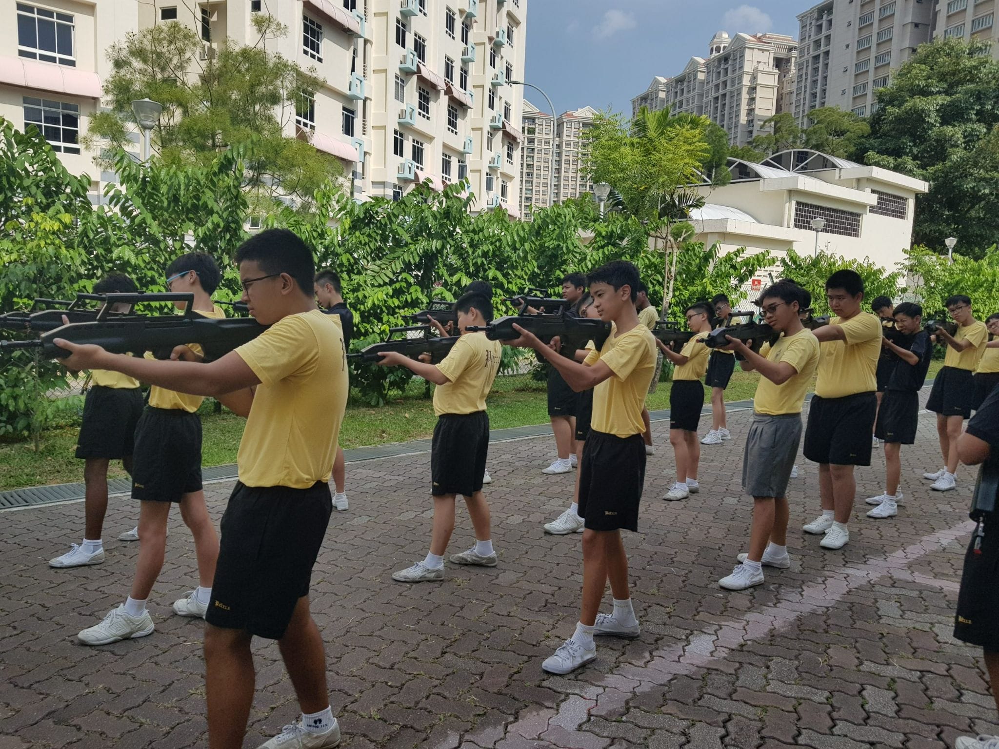 Young students in yellow shirts and black shorts stand in rows outdoors, aiming rifles during a drill exercise.