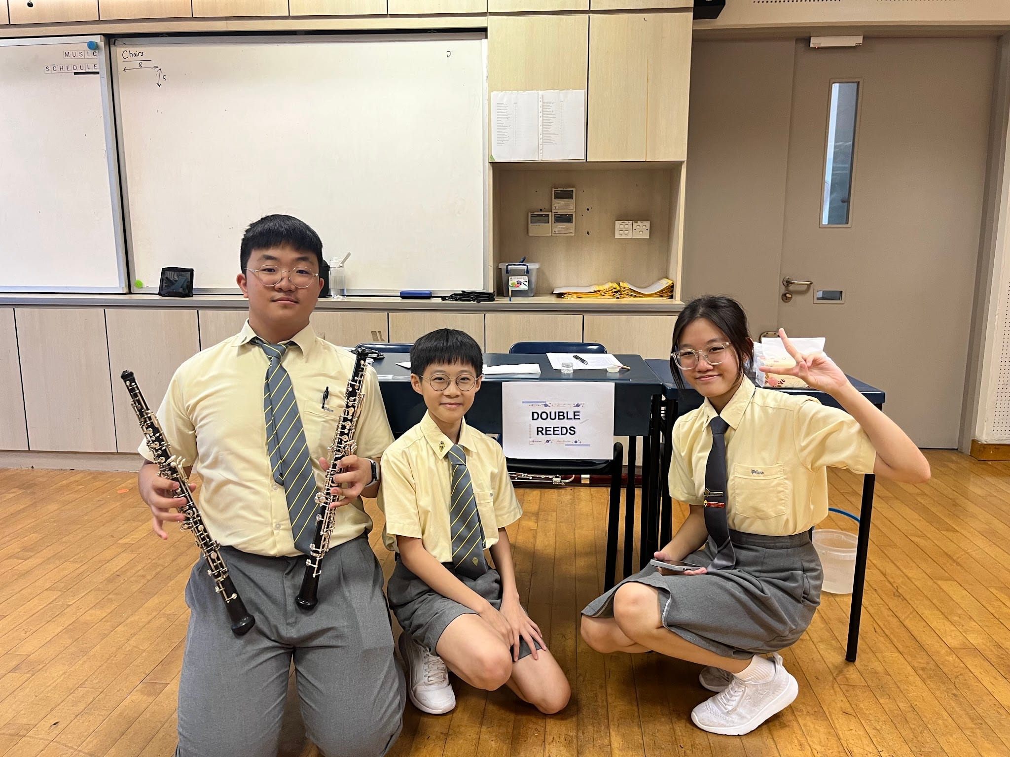 Three school students in uniform pose near a Double Reeds sign, two holding oboes in a music classroom.