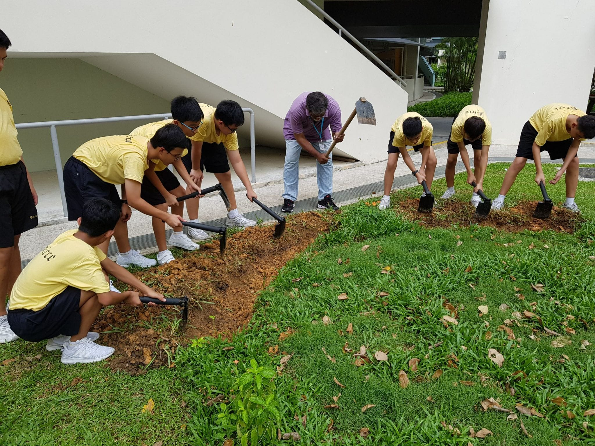 Students in yellow shirts and an adult dig soil on a grassy area beside a building using shovels and rakes.