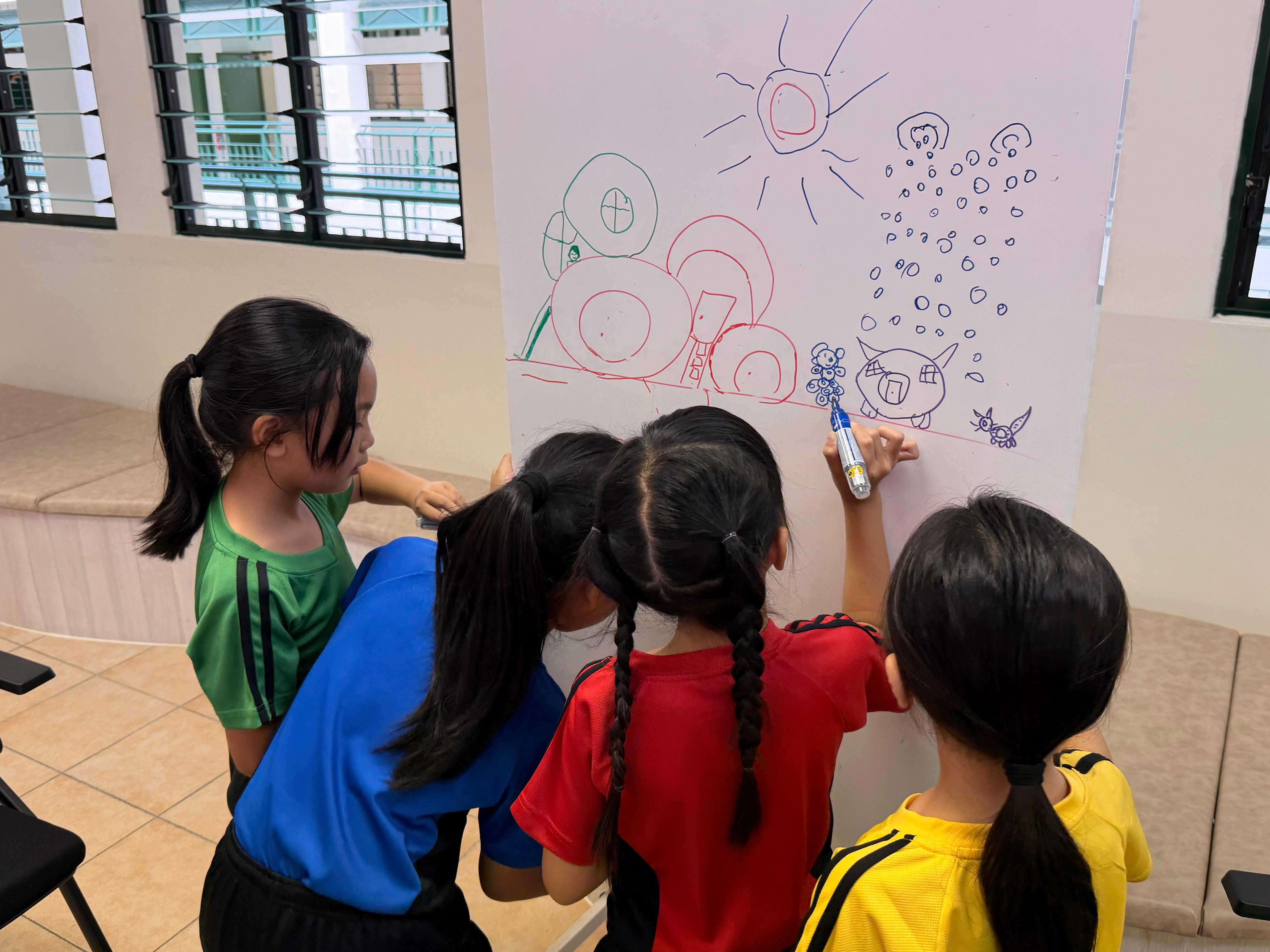 Four children drawing colorful shapes and figures together on a large white board with markers.