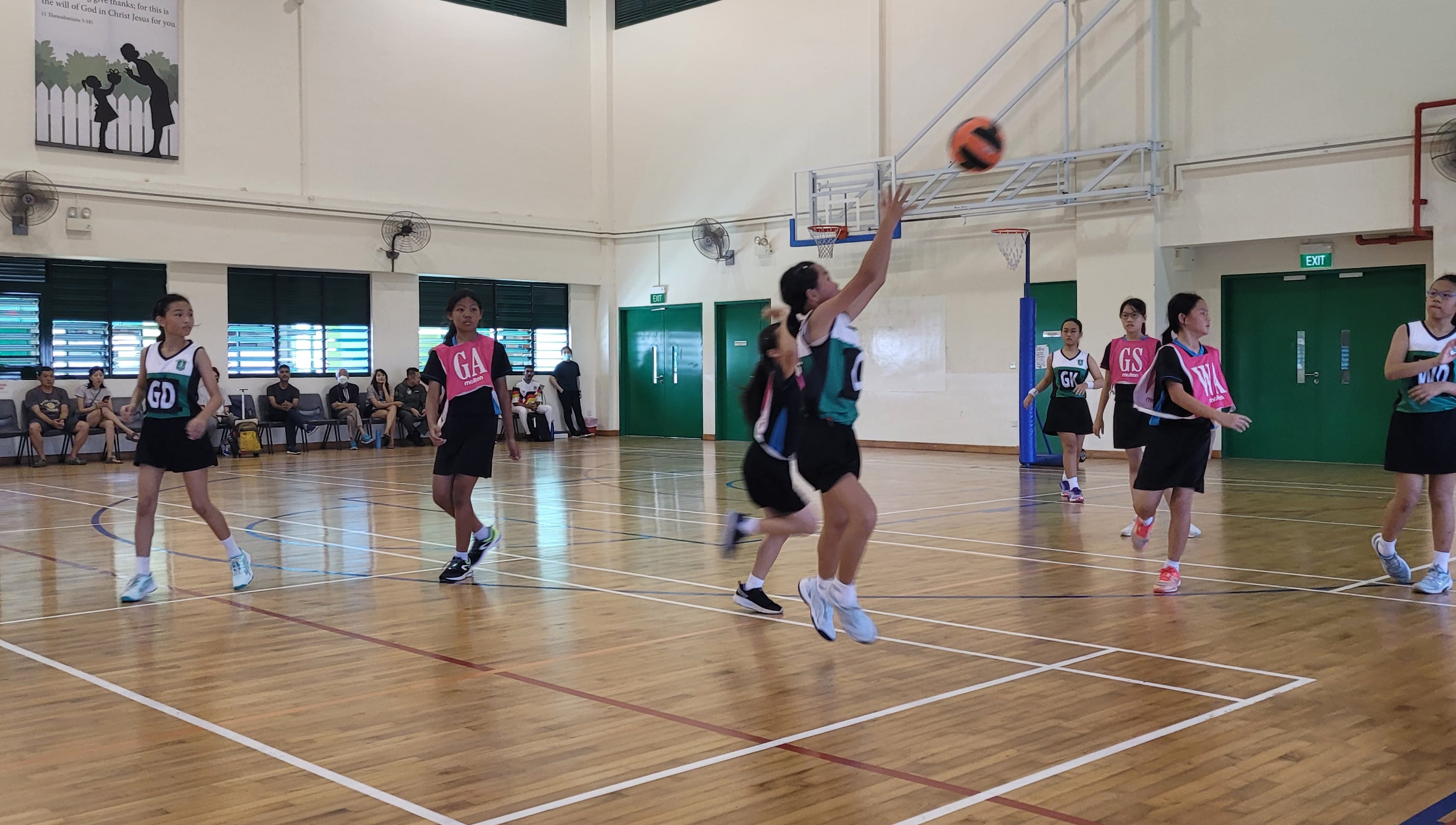 Girls in pink and black bibs play netball indoors, with one player shooting the ball toward the hoop.