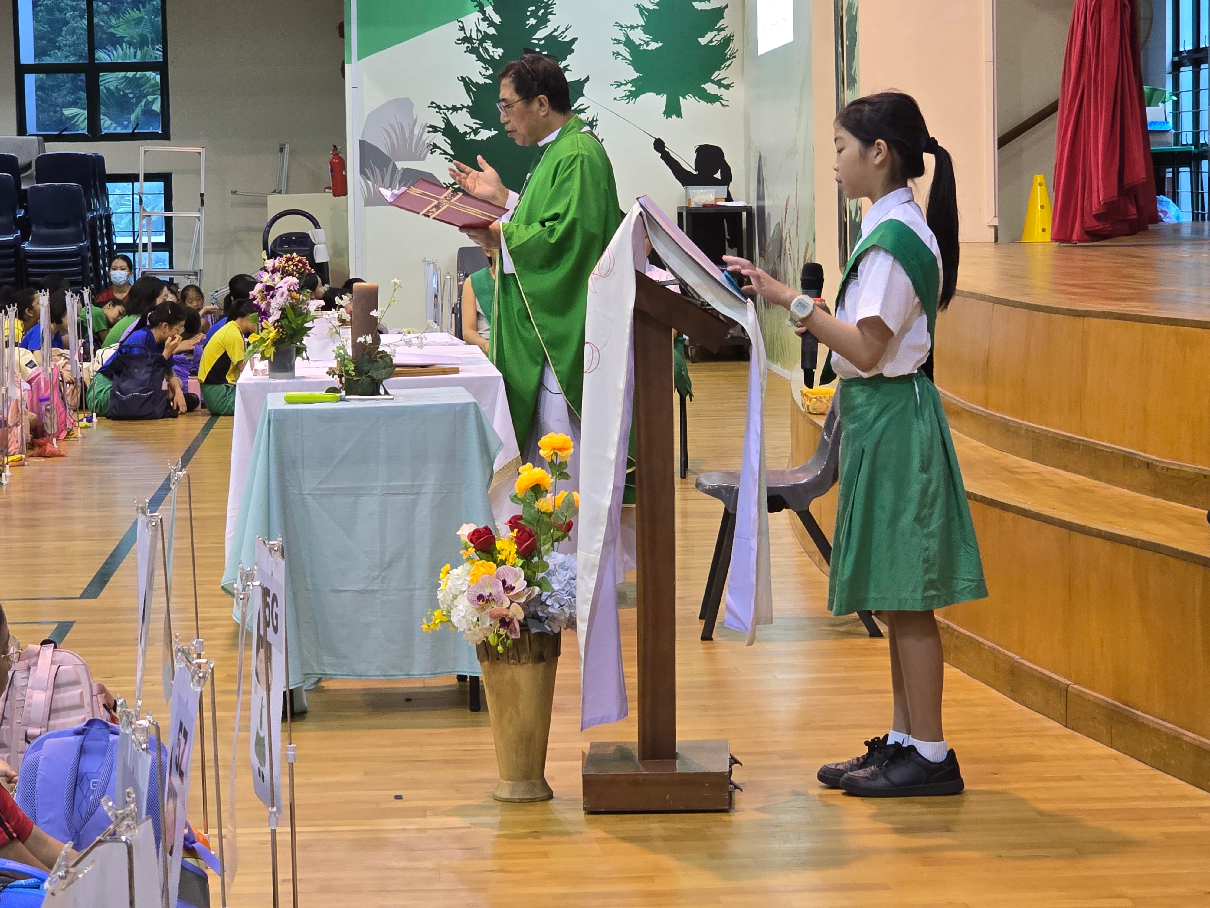 A priest in green vestments reads from a book at a podium while a girl in a green school uniform stands nearby.