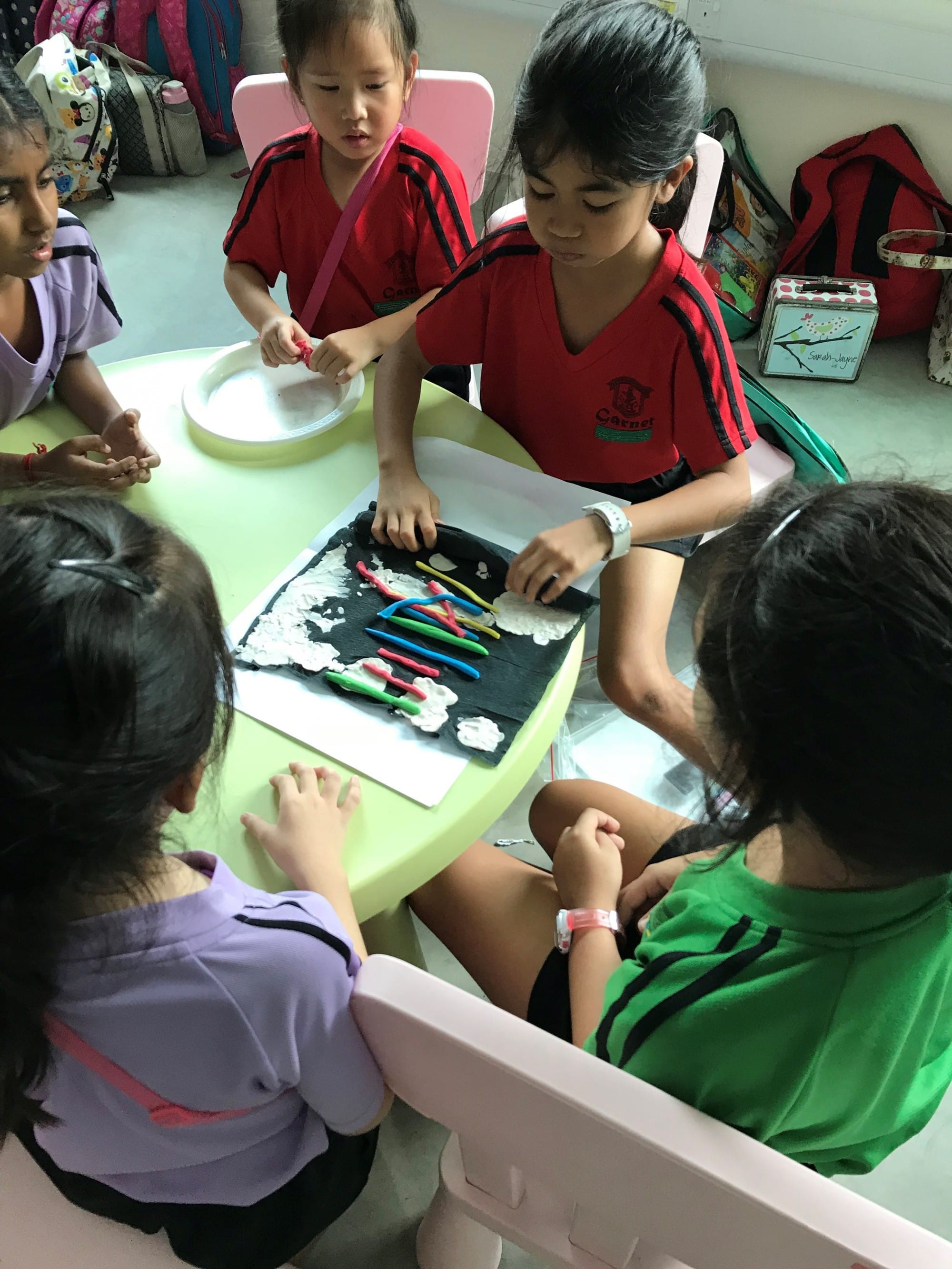 Four young girls in school uniforms sit around a table working on a craft with colorful clay strips on black paper.