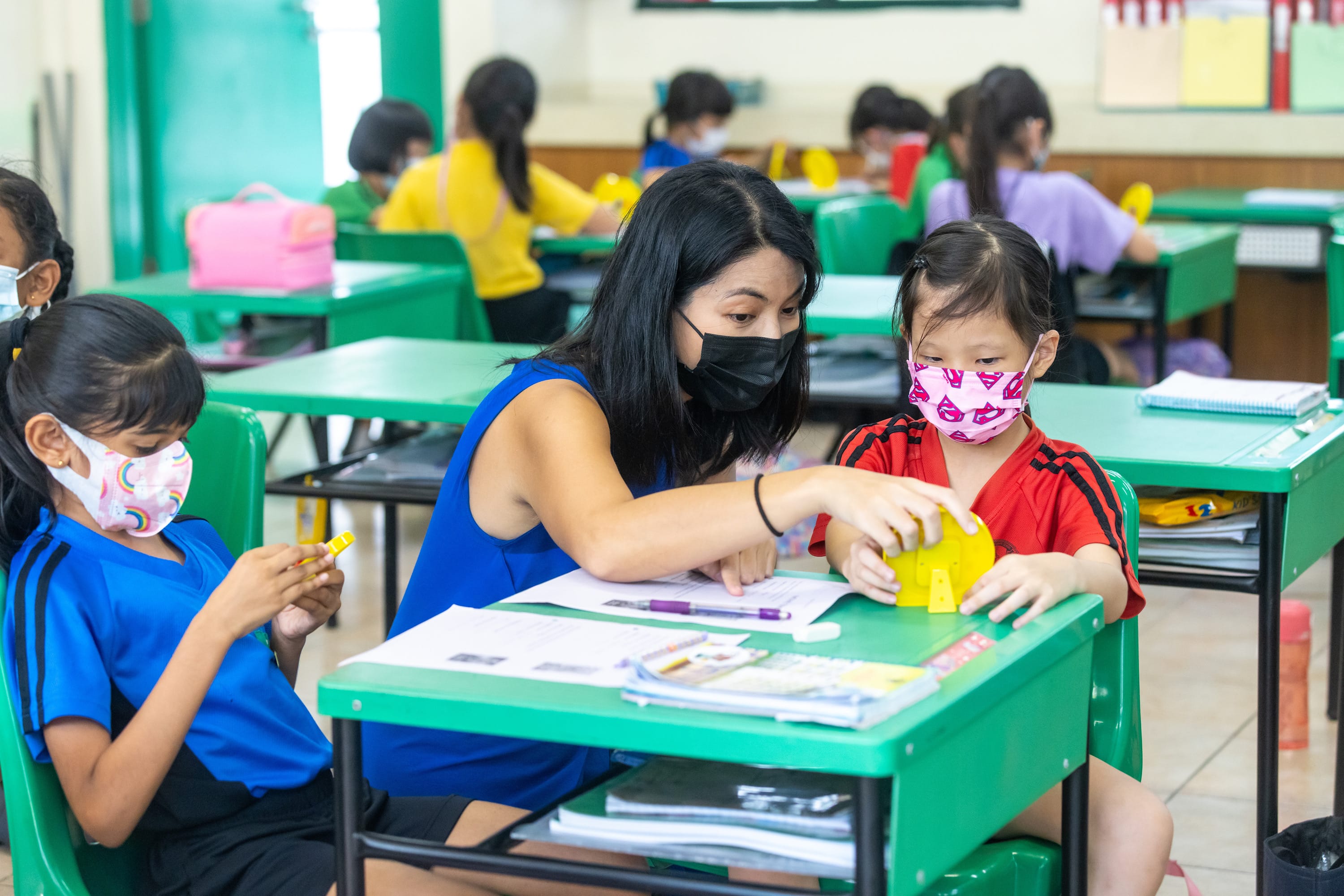 A masked teacher helps a young masked child with a yellow object at a green desk in a classroom full of students.