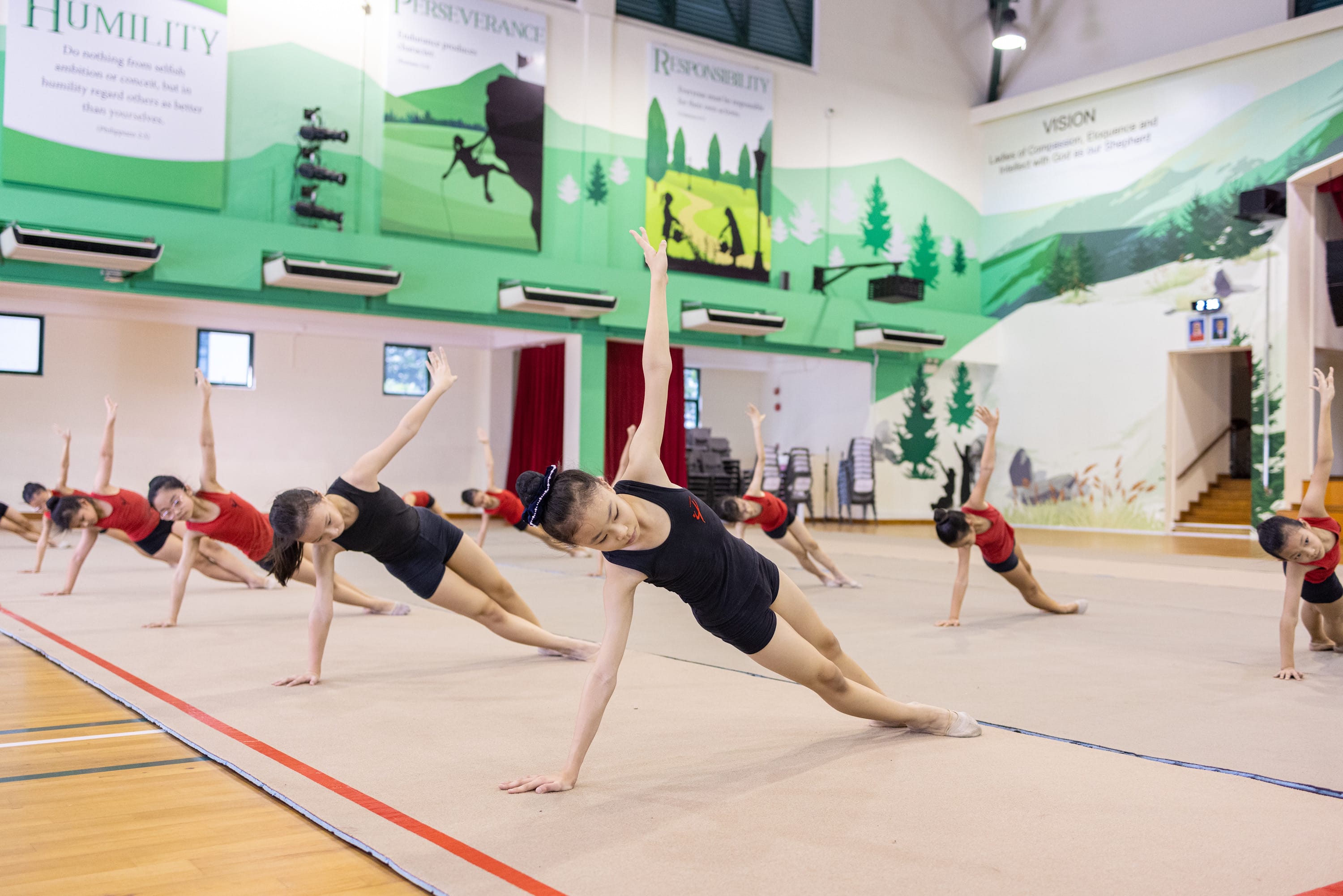 Young gymnasts in red and black leotards perform side stretch poses on a mat in a decorated school gymnasium.
