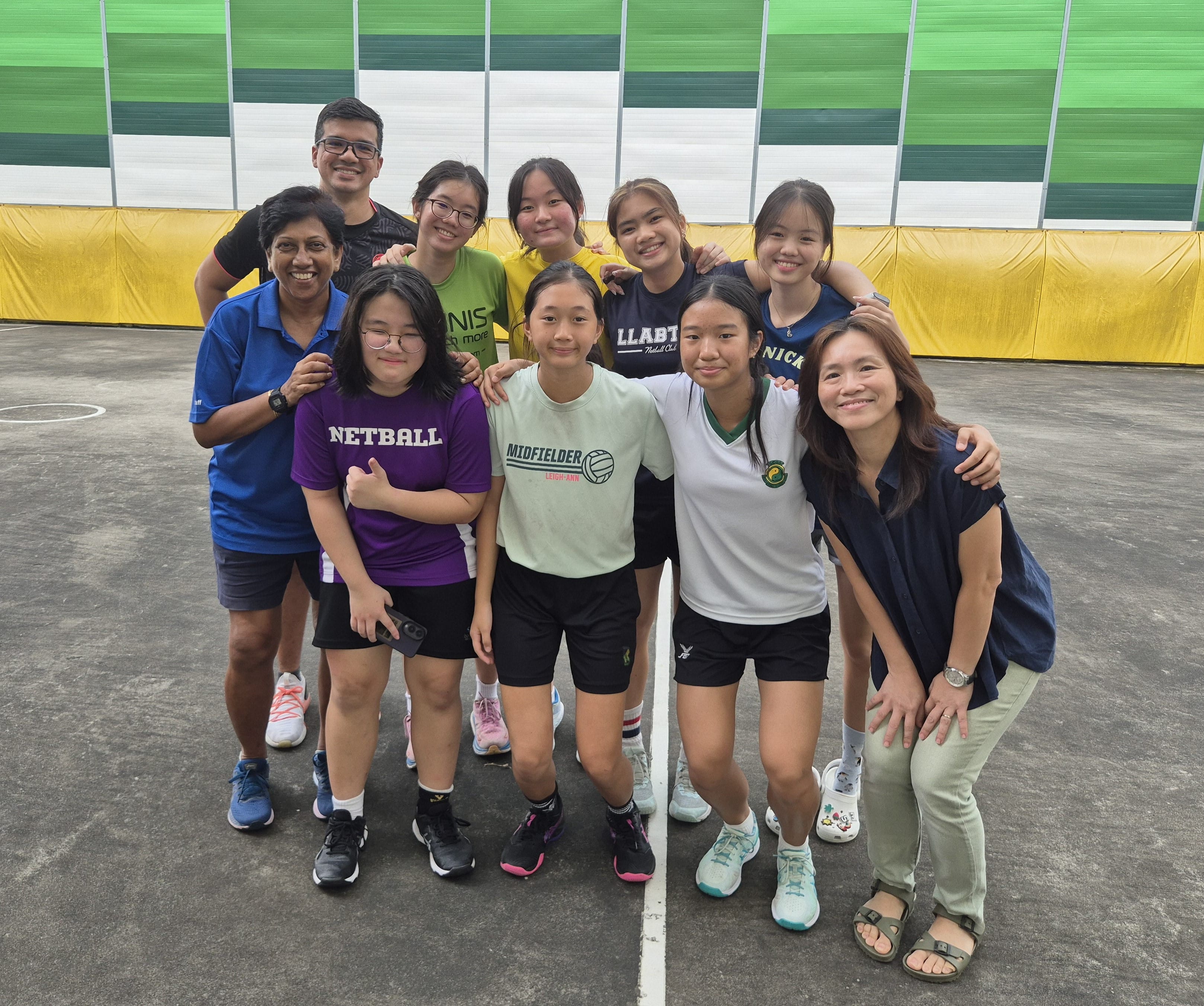 A group of ten people, mostly young girls in netball jerseys, pose smiling on an outdoor court.