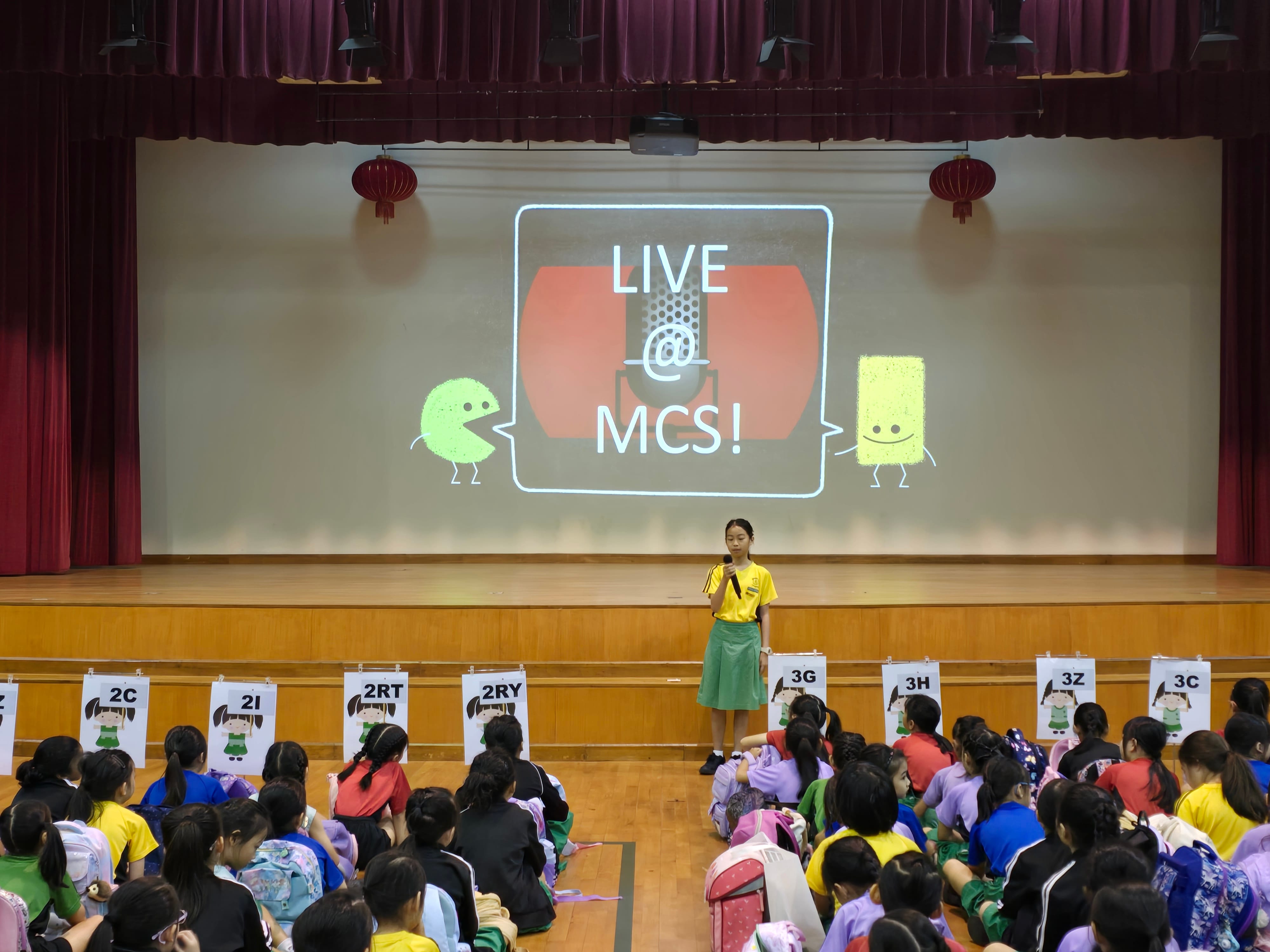 A girl holds a microphone on a school stage with a projected screen reading "LIVE @ MCS!" while students sit on the floor.