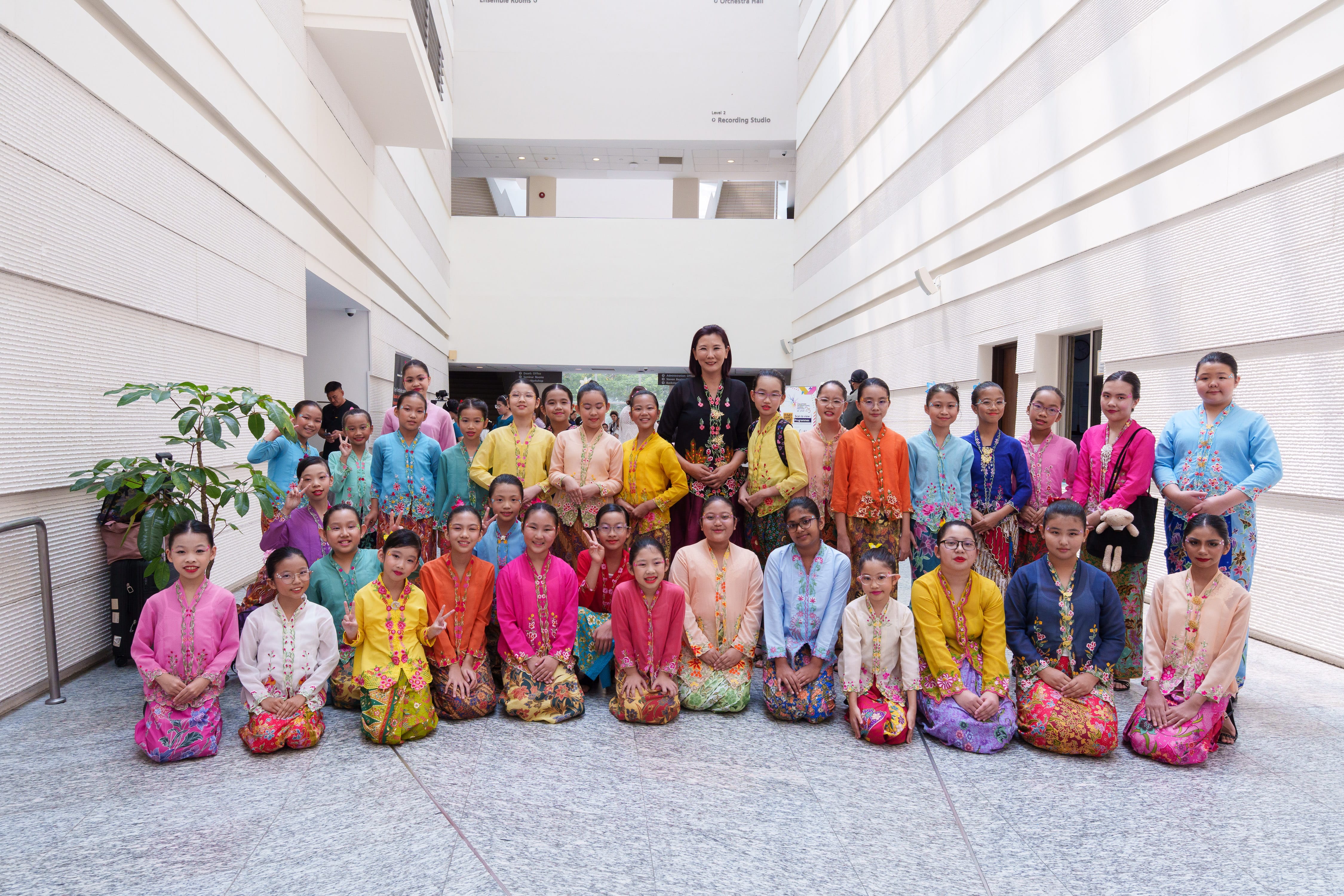 A large group of girls in colorful traditional Southeast Asian costumes pose with an adult woman in an indoor atrium.
