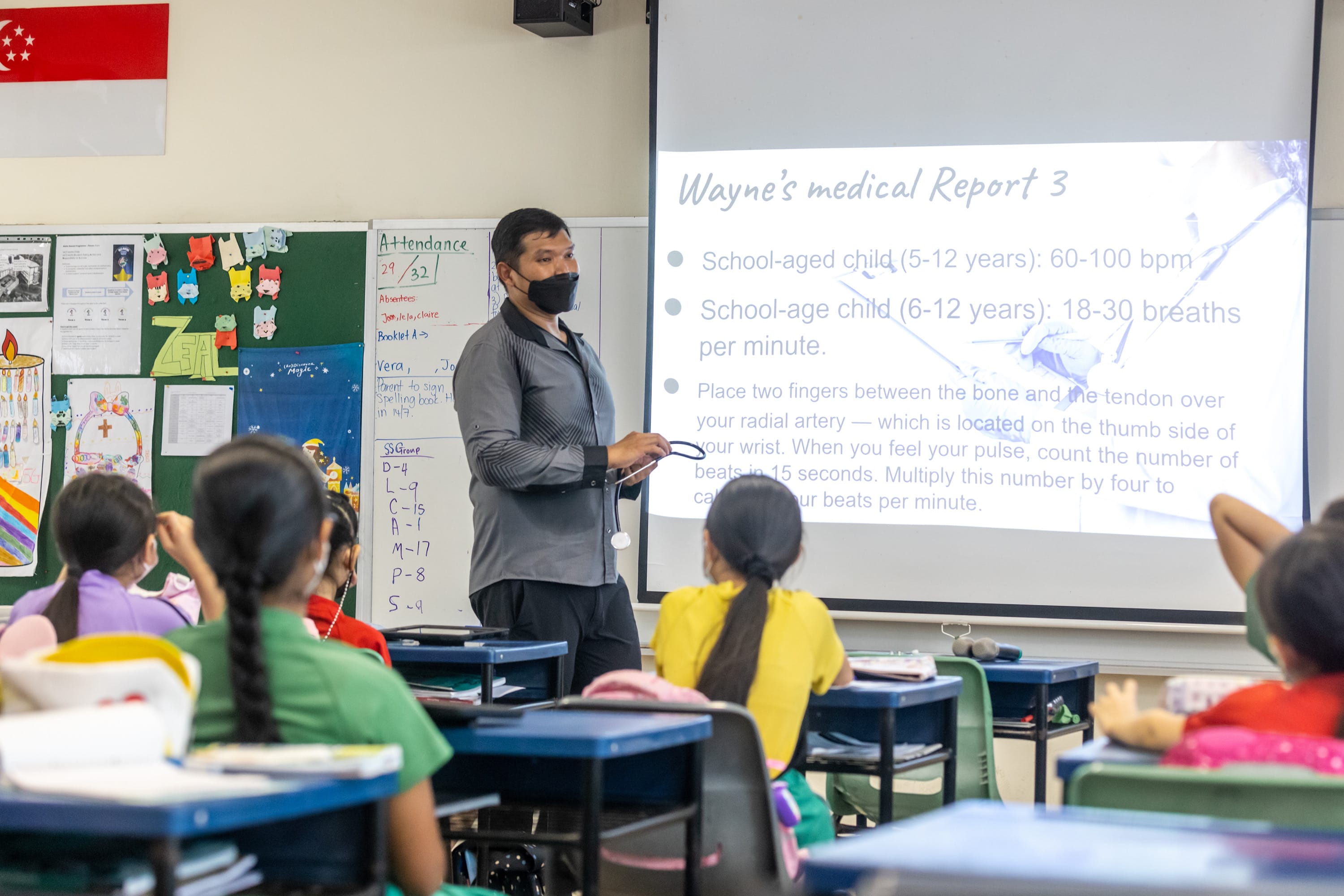 A masked teacher presents a medical slideshow to young students seated at desks in a colorful classroom.