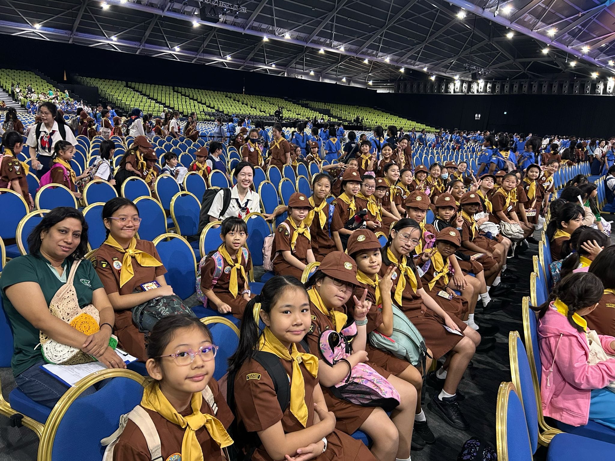 Large group of children in brown scout uniforms with yellow scarves seated in a large indoor arena with blue chairs.