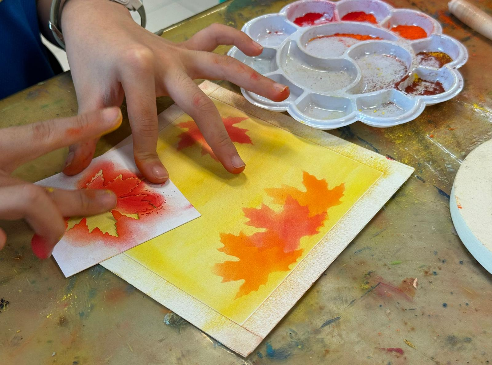 Child's hands creating fall leaf art using stencils with orange and red paint on yellow watercolor paper.