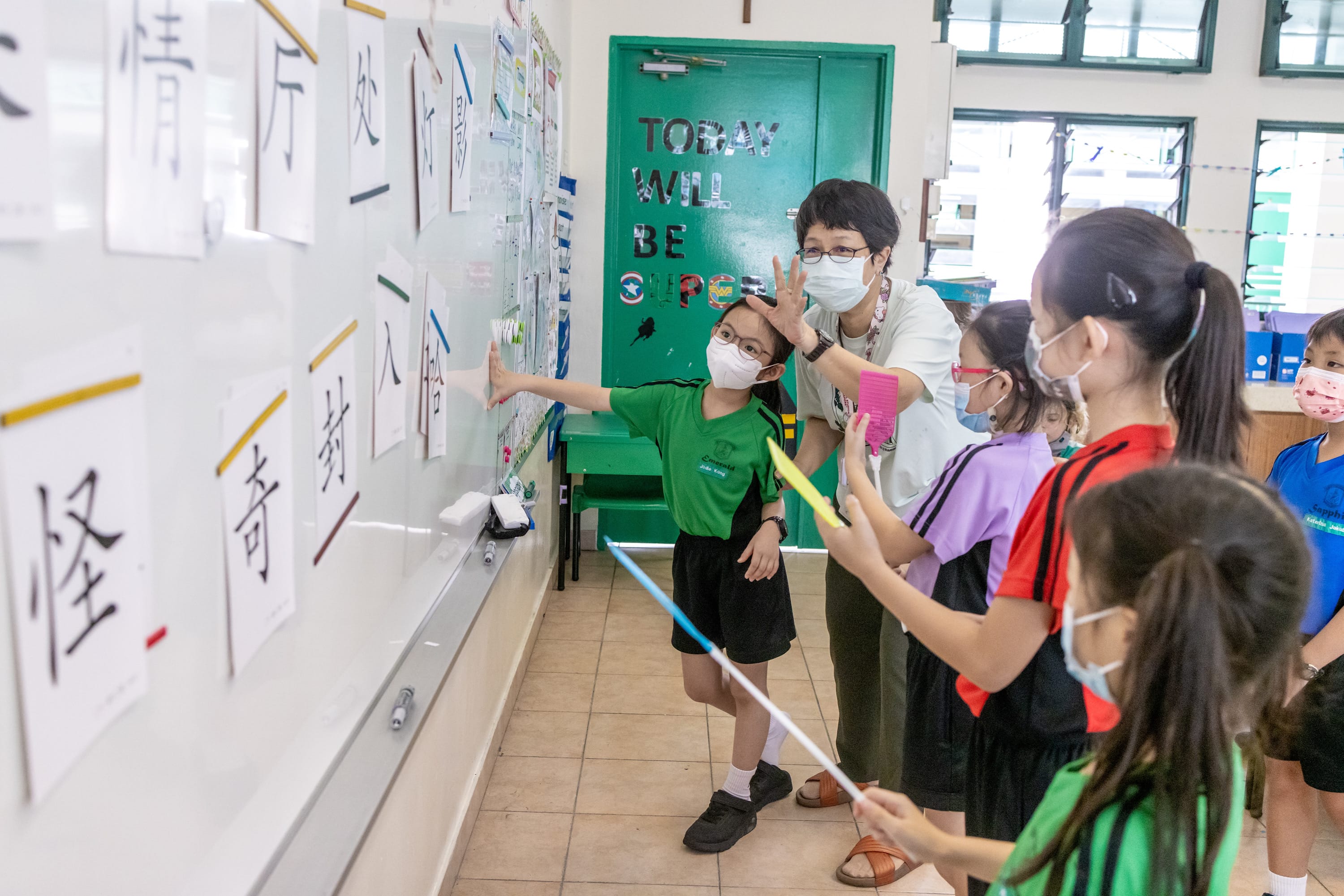 A masked teacher and young students point at Chinese character flashcards on a whiteboard in a classroom.