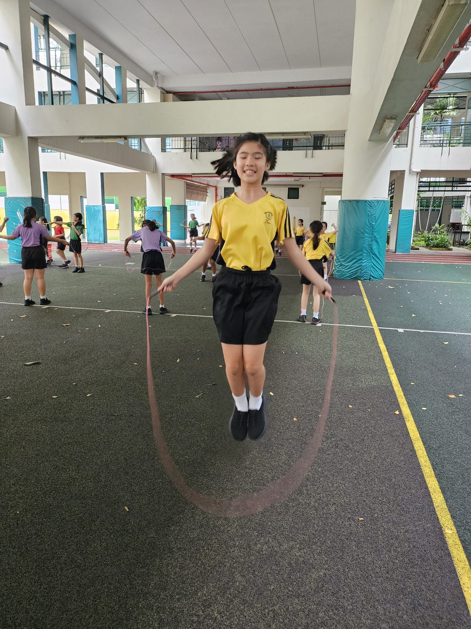 A smiling girl in a yellow shirt jumps rope mid-air in a school sports hall with other students in the background.