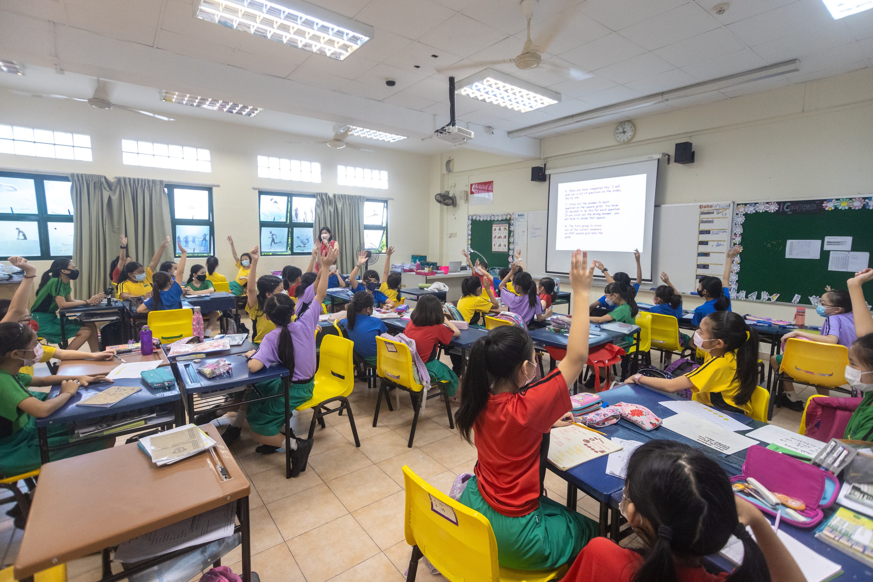 Students in colorful uniforms raise hands at blue desks while a teacher stands near a projector screen.