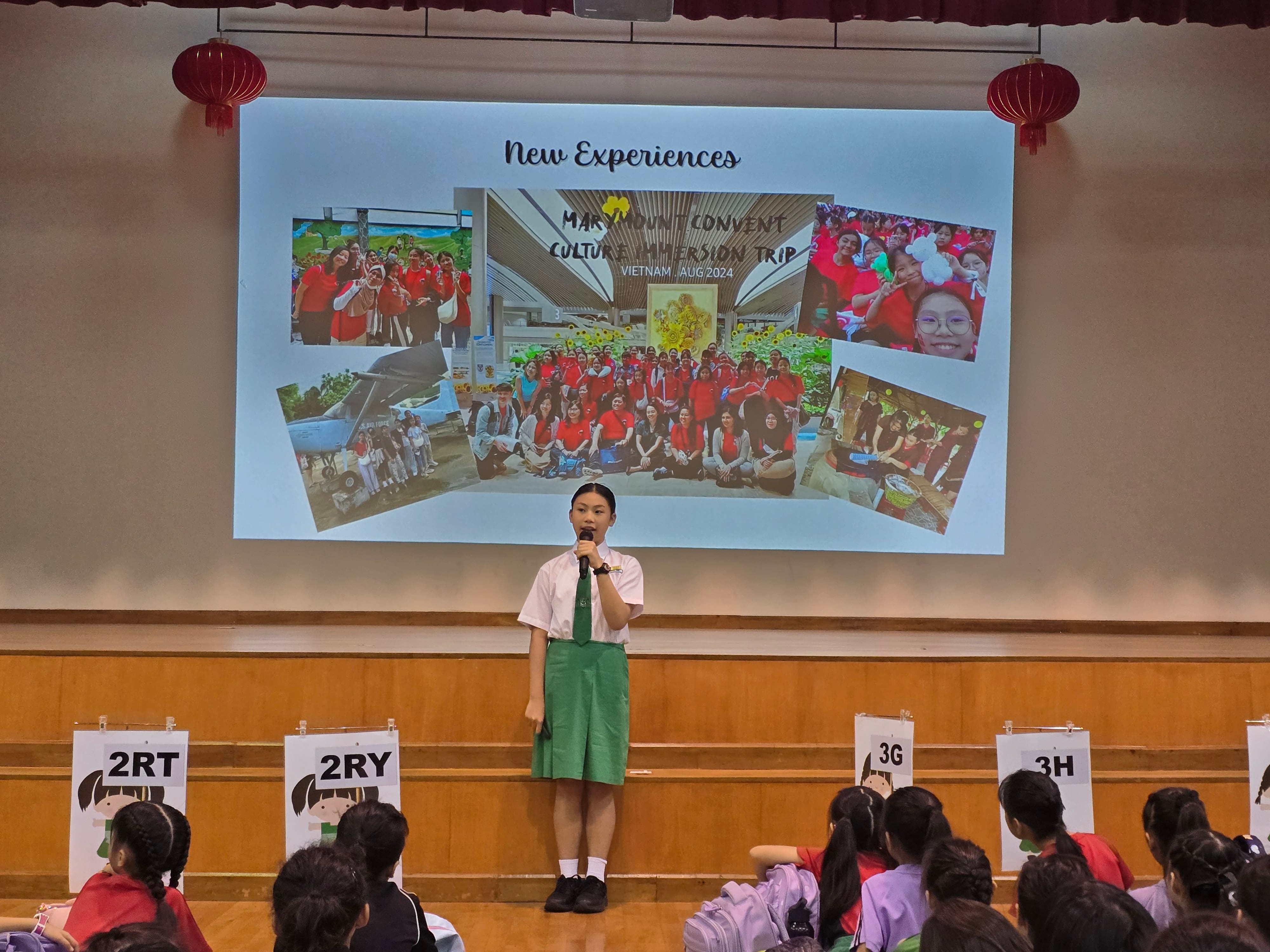 A student in uniform speaks into a microphone on stage before a projected slide about a Vietnam culture immersion trip.