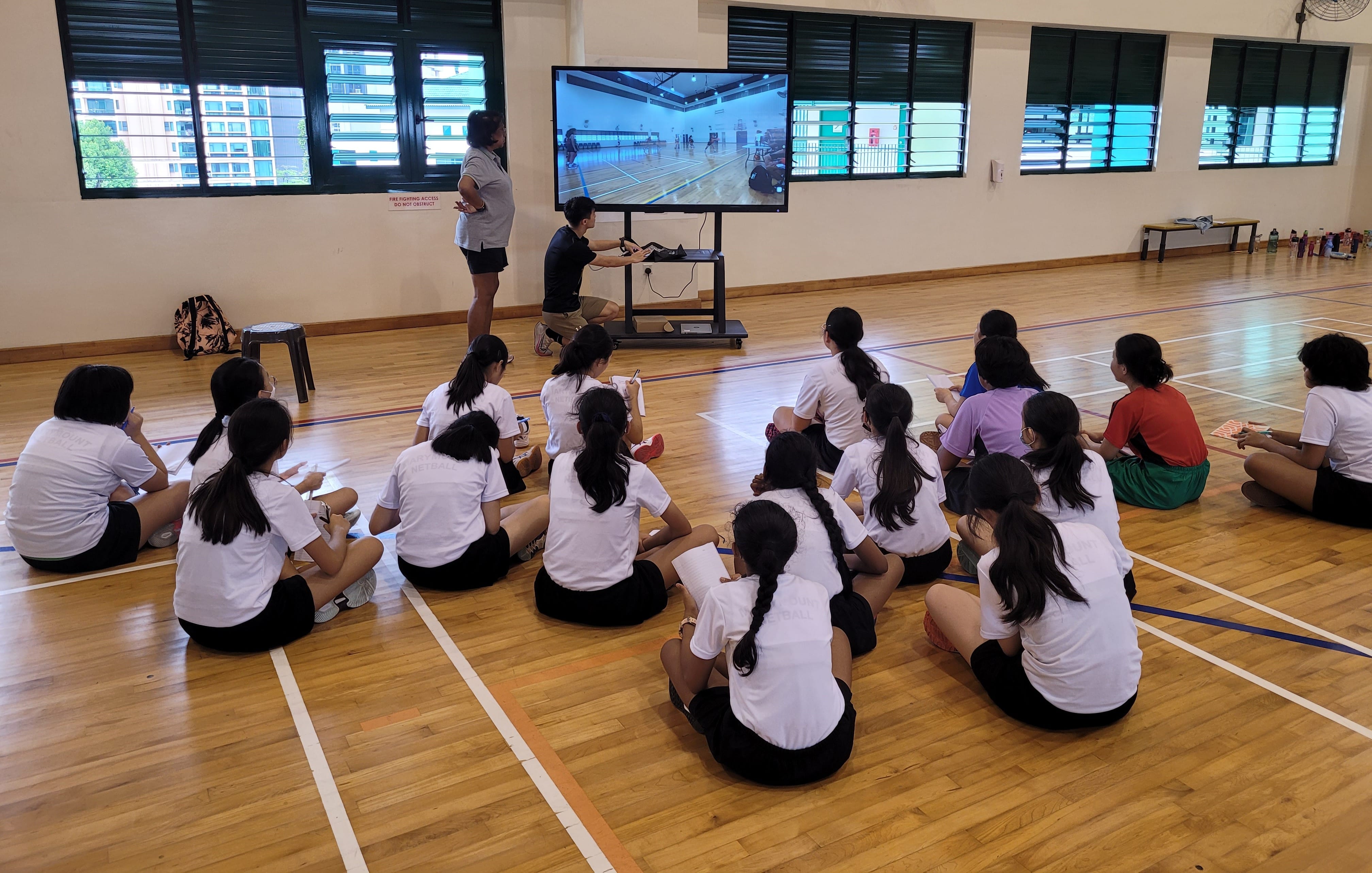 Students in white shirts sit on a gym floor watching a large screen showing a sports court presentation.