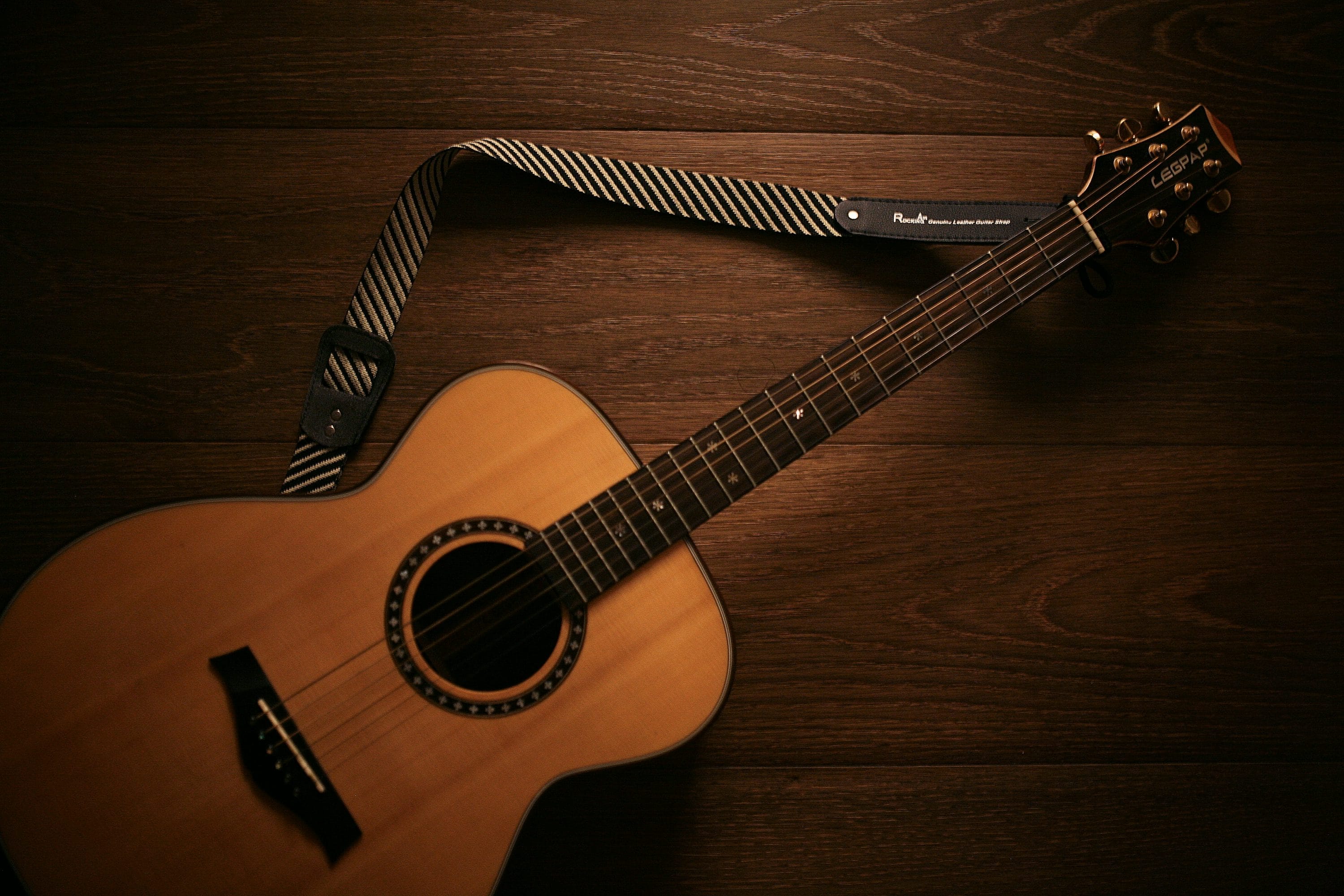 Acoustic guitar with a striped strap lying on a dark wooden floor, viewed from above.