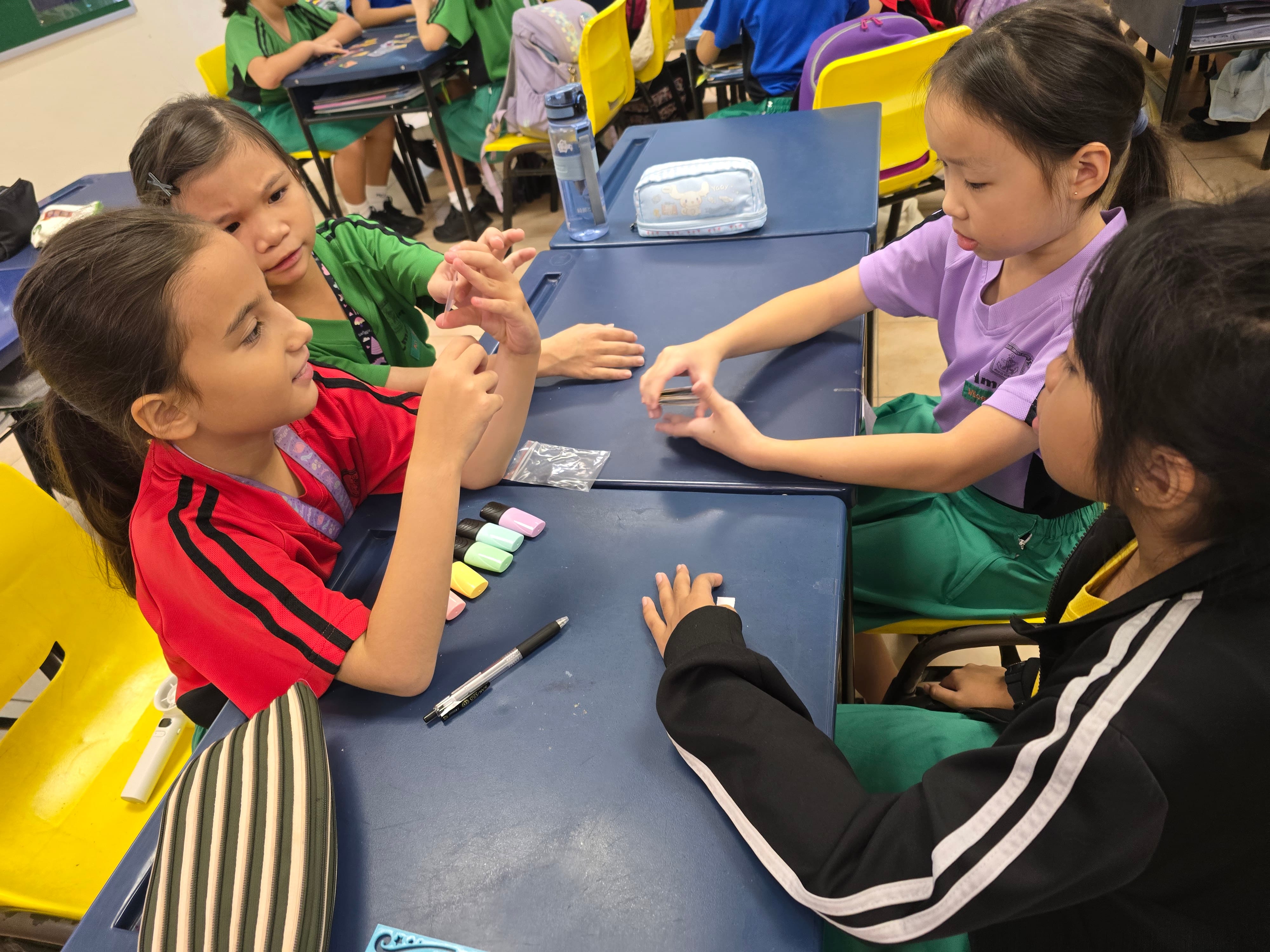 Four young girls sit around a blue desk in a classroom, handling small colorful erasers and stationery.