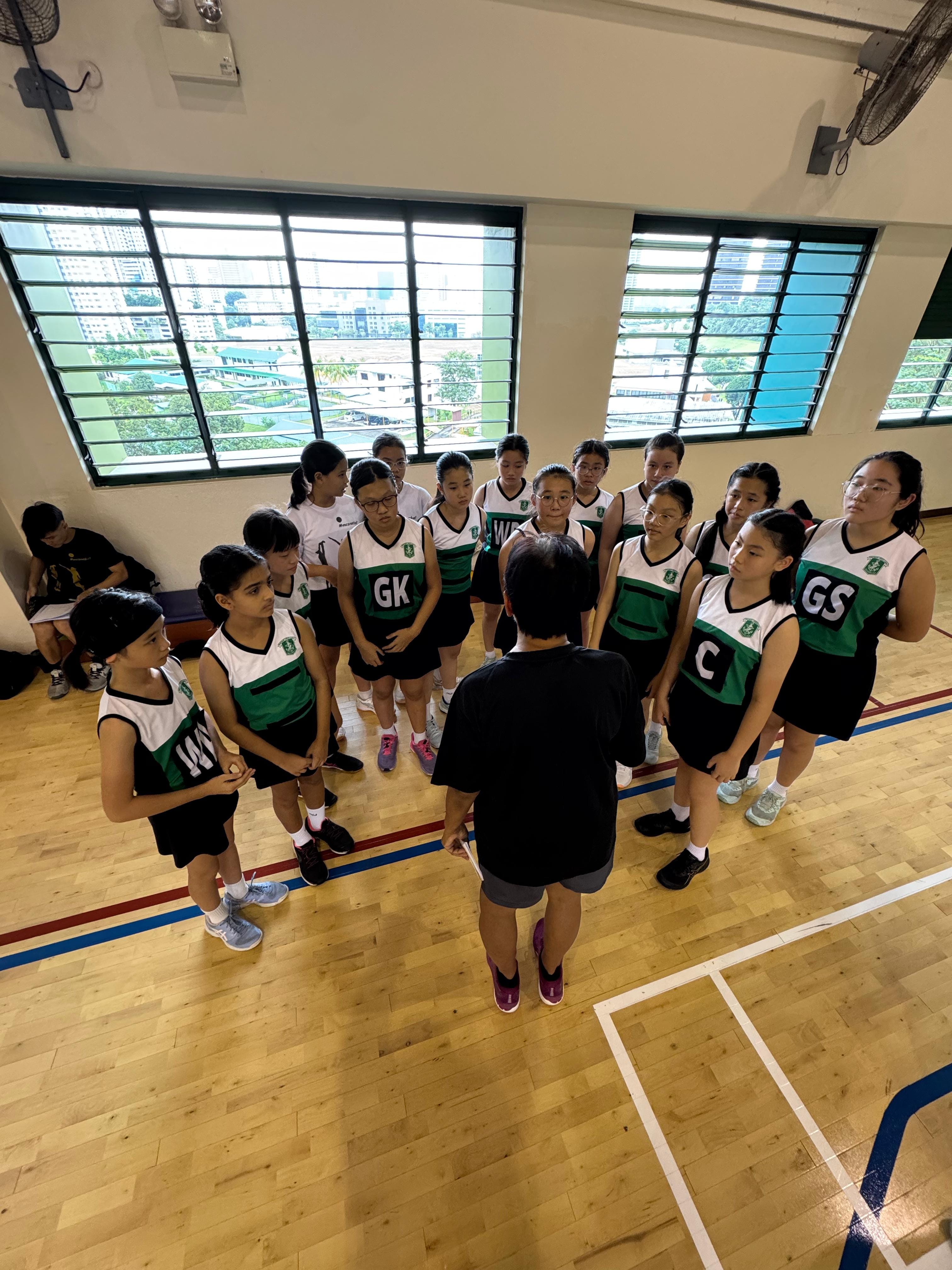 A coach addresses a group of young girls in green and white netball uniforms gathered in a circle on an indoor court.