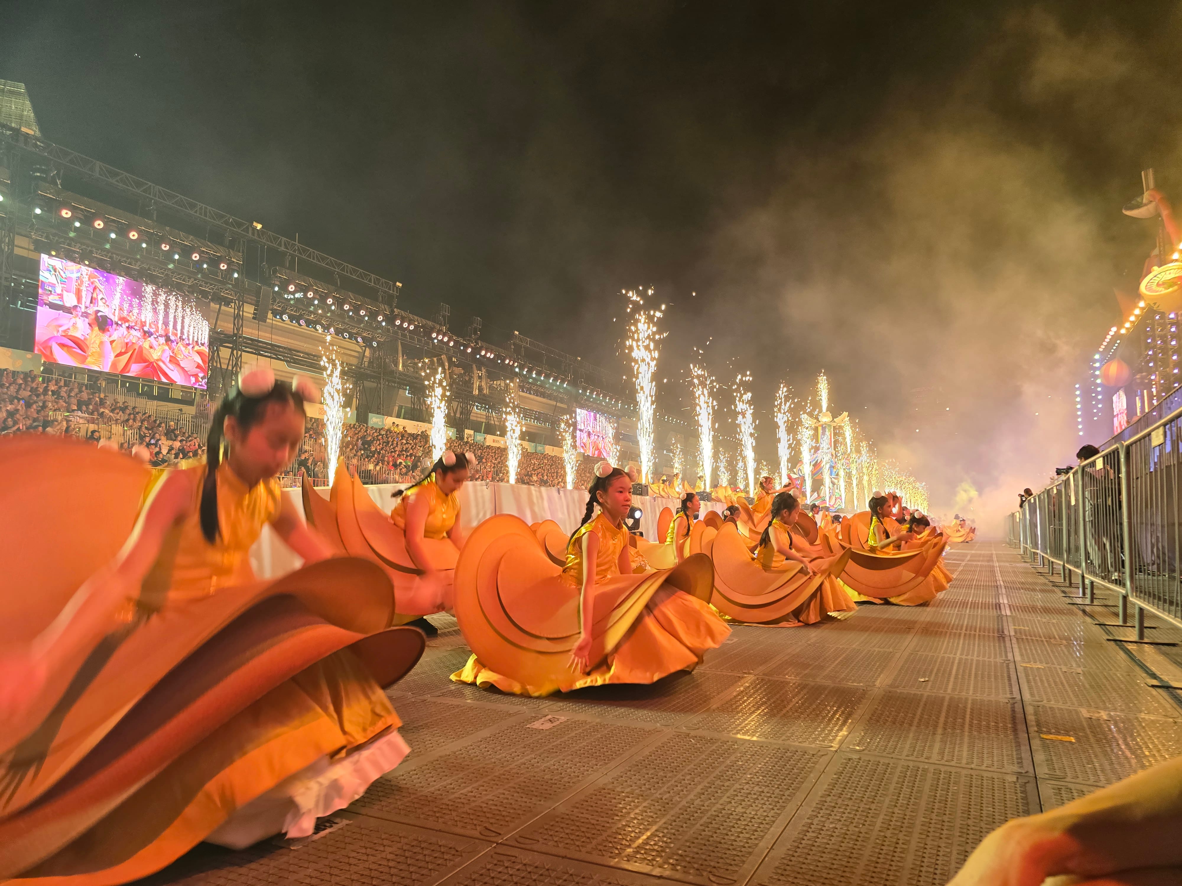 Performers in golden gowns dance along a parade route at night with pyrotechnic fountains blazing behind them.