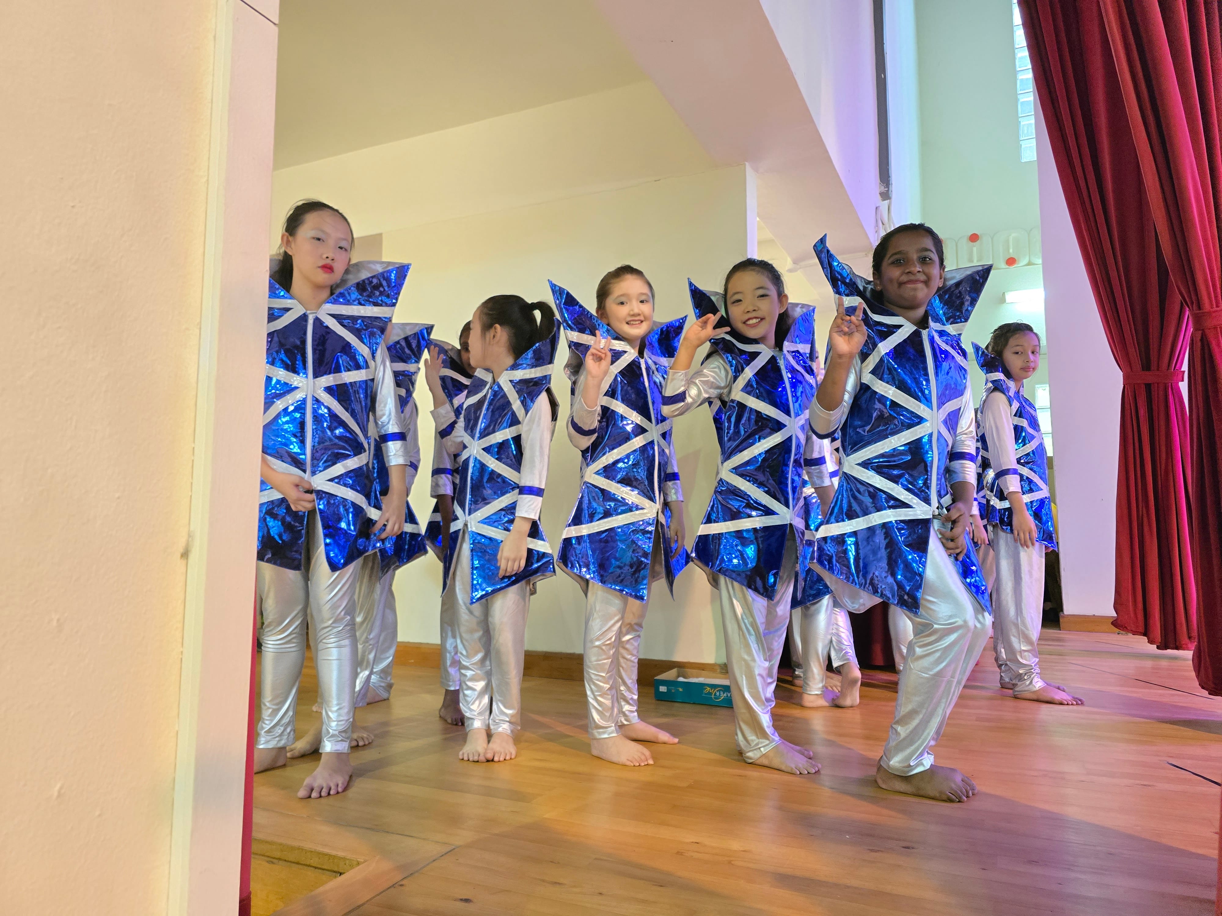 Five girls in shiny blue and silver geometric costumes pose barefoot on a wooden stage near a red curtain.