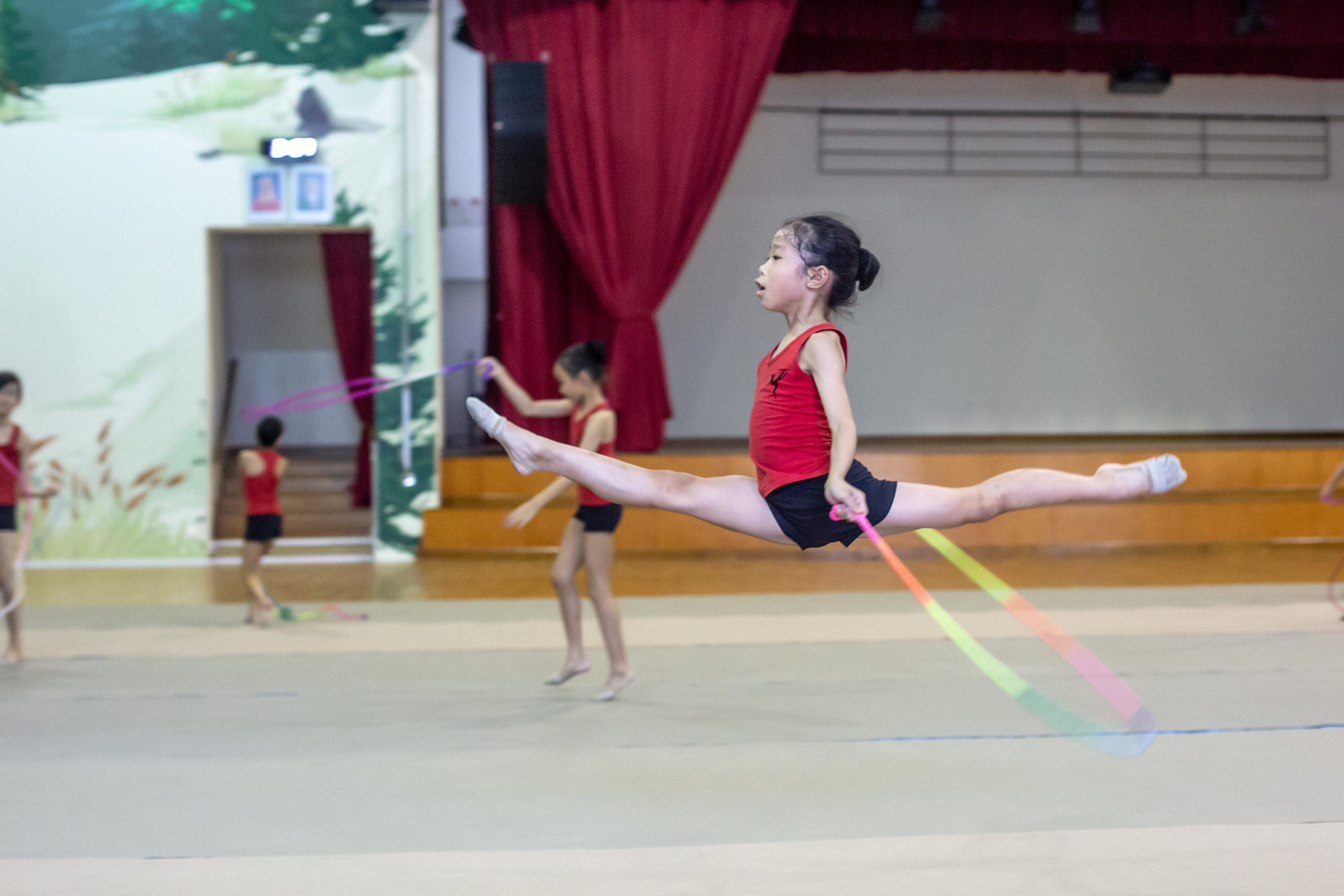 Young girl in red leotard performs a mid-air split leap holding a colorful ribbon during rhythmic gymnastics.