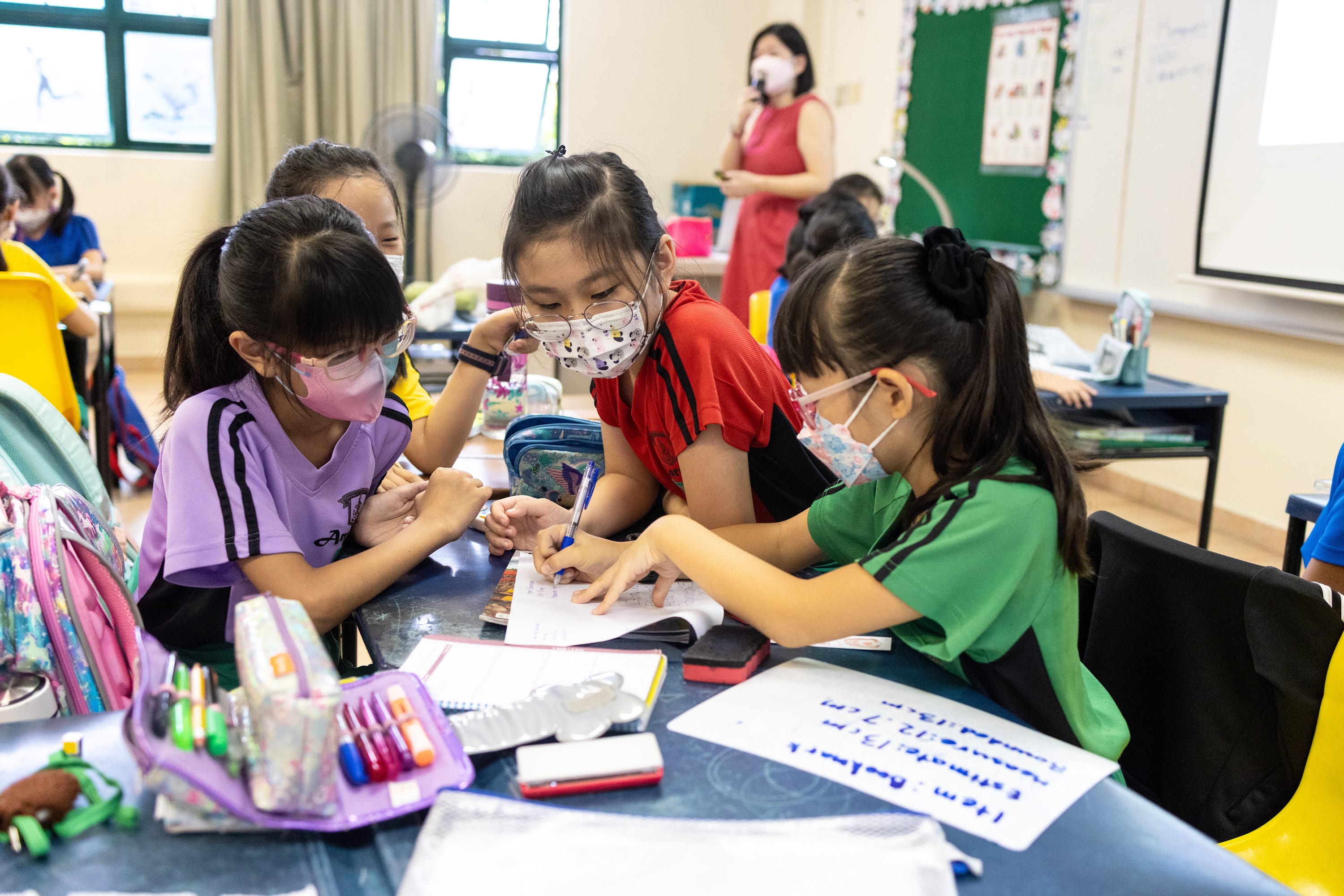Three masked young girls collaborate over a notebook at a school desk, with a teacher standing in the background.