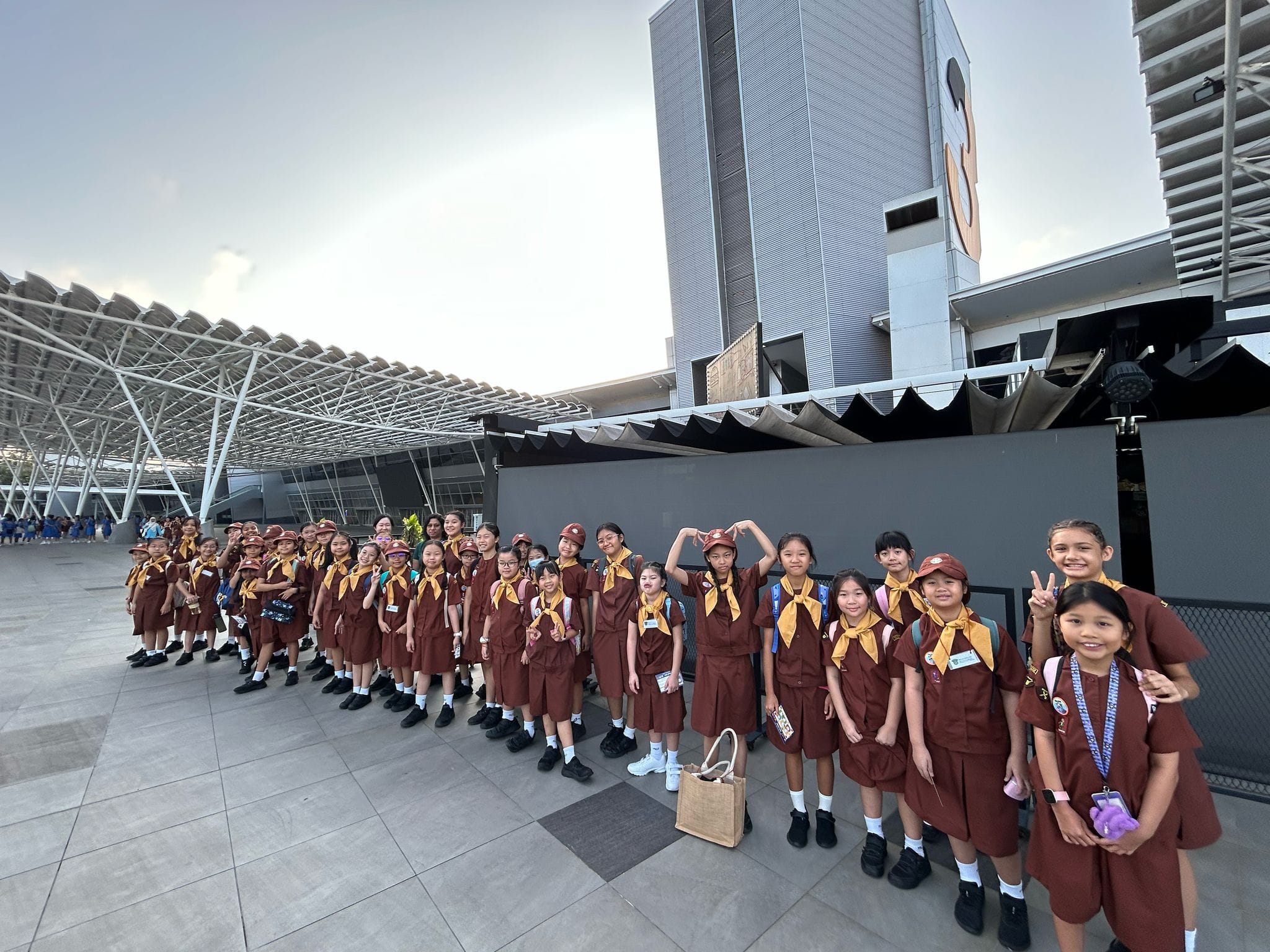 A large group of girls in matching brown uniforms and yellow scarves pose outdoors in front of a modern building.