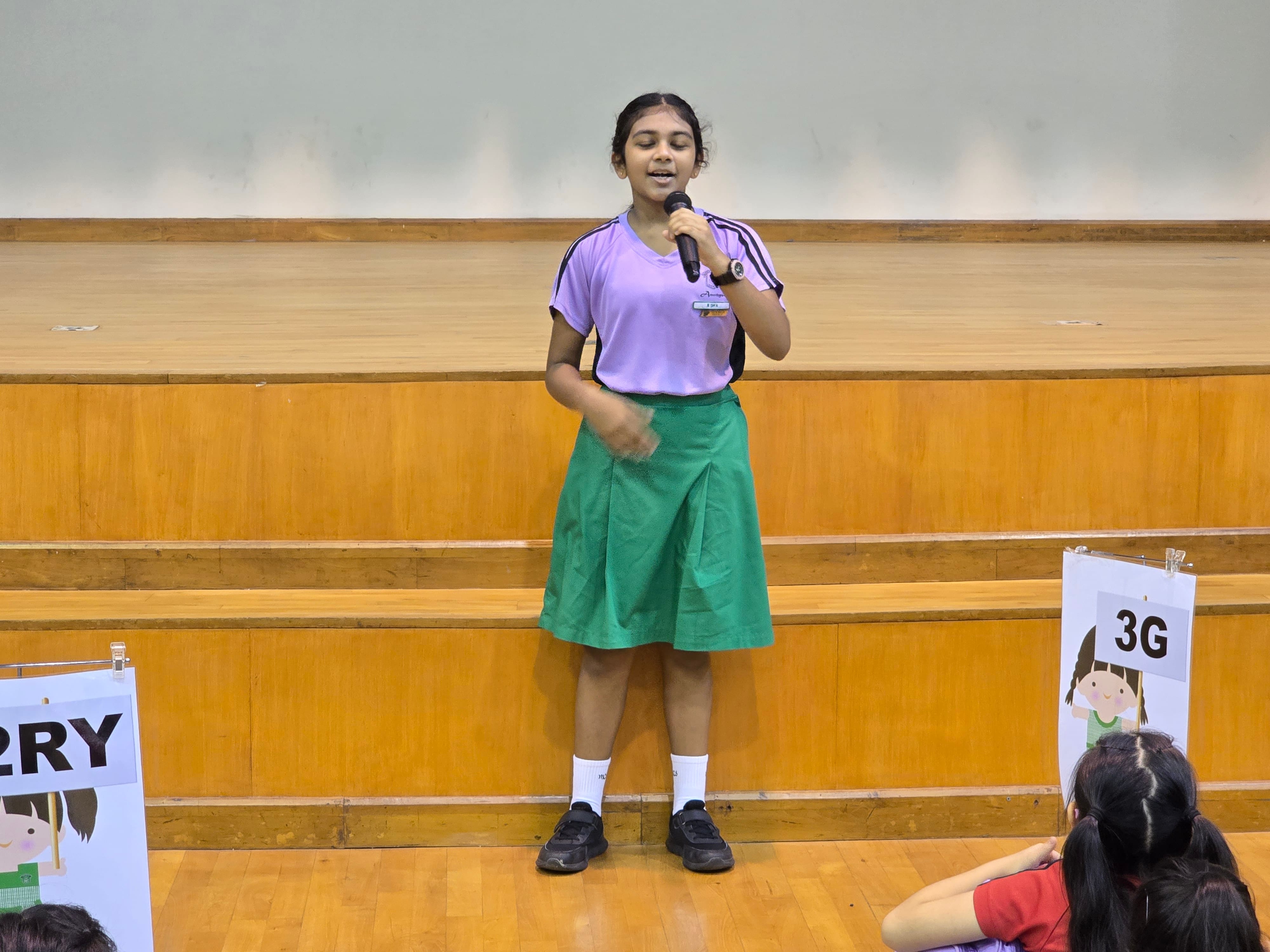 A young girl in a purple top and green skirt speaks into a microphone on a school hall floor.
