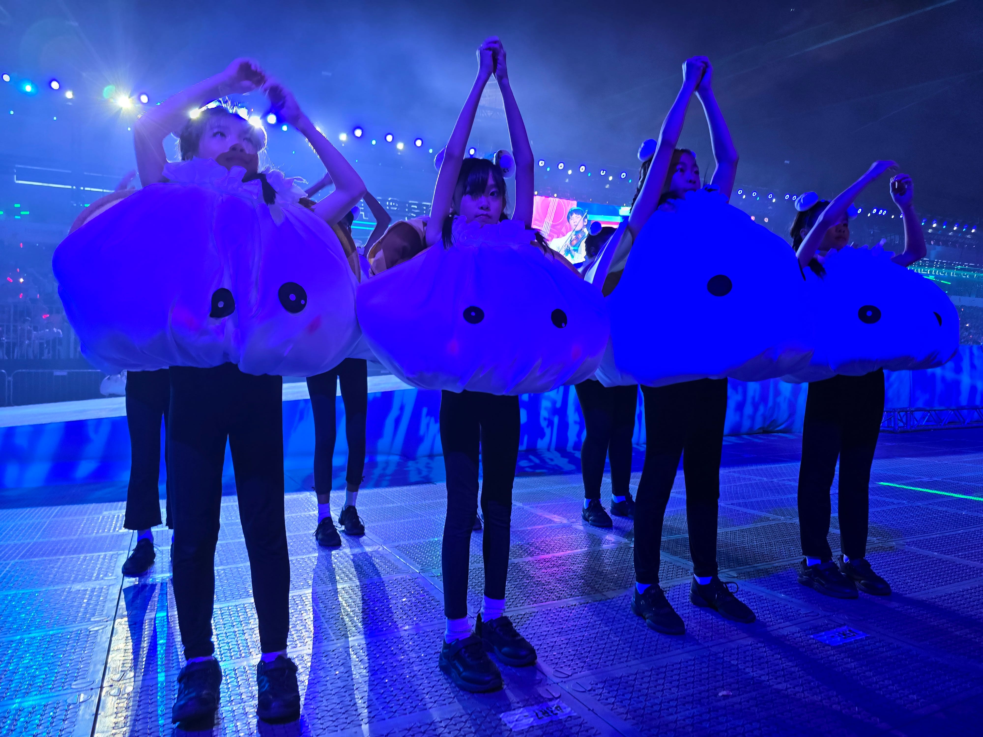 Performers in blue round cartoon creature costumes raise their arms on a stage lit with blue lights.
