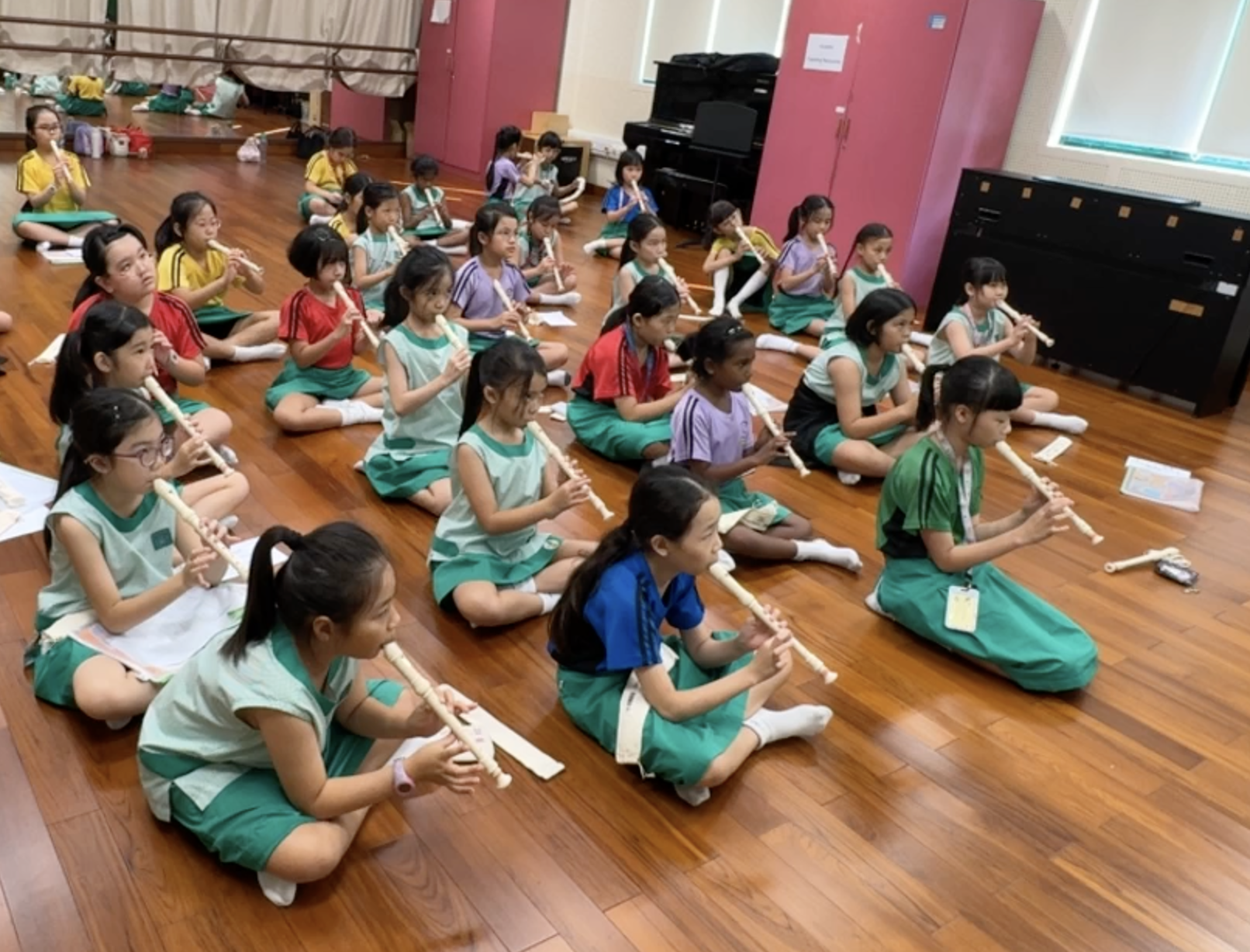 A large group of children in green uniforms sit on a wooden floor playing recorders in a music room.