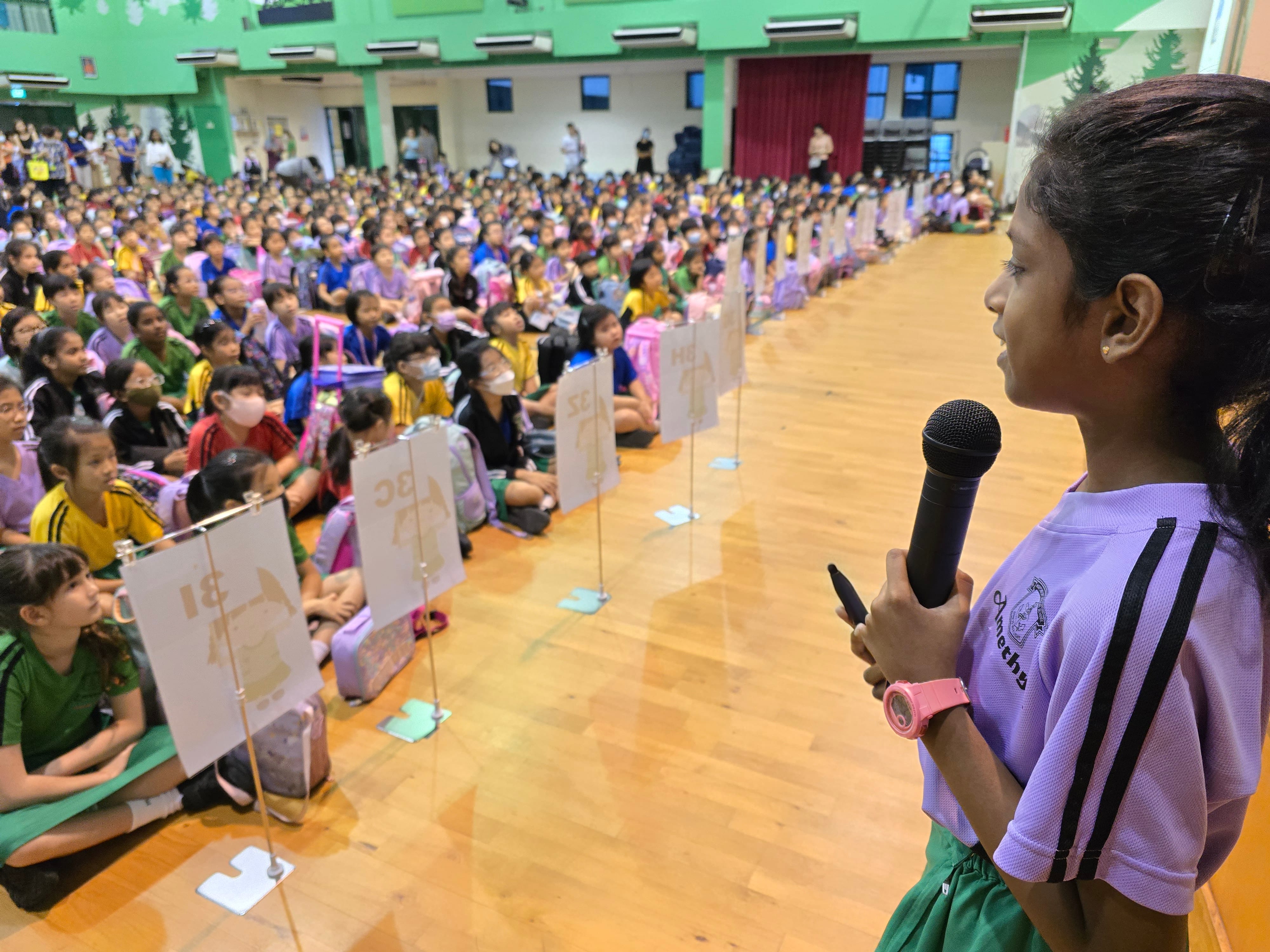 A young girl in a purple school uniform holds a microphone, addressing a large crowd of students seated on a gym floor.